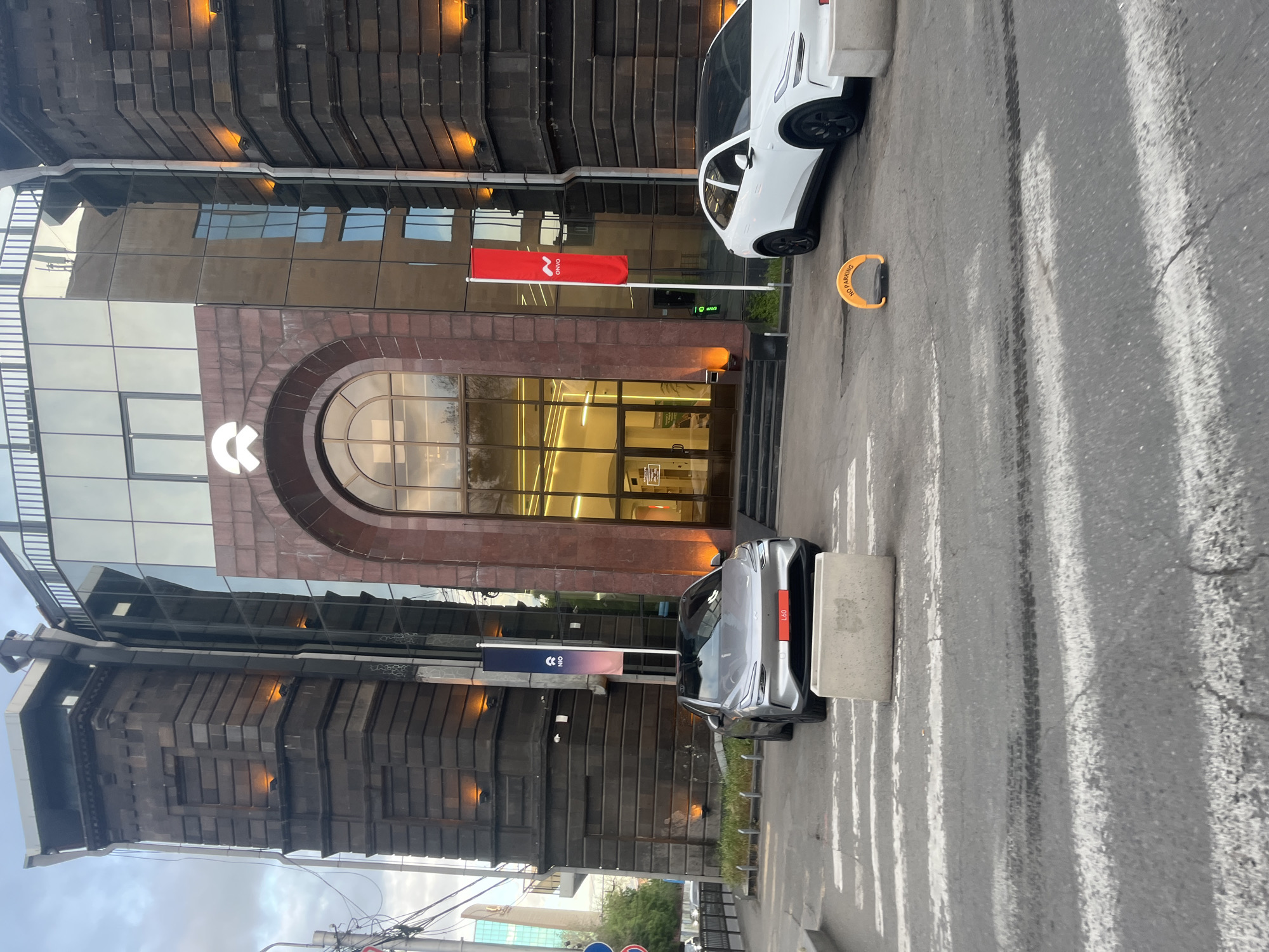 The image shows the exterior of a modern building in Yerevan, Armenia. The building is multi-story, with a facade that combines glass and brown stone elements. There is a large archway above the entrance. A white logo, possibly for a company, is visible on the facade. Two cars, one white and one silver, are parked on the street in front of the building. A red flag with a white logo is flying near the entrance. A blue flag with a company logo is also visible on the left side of the building. Street markings and a traffic barrier are visible on the street. The buildings surrounding are a mix of old and new architectural styles, suggesting the area is an urban environment with a blend of historical and modern development. Power lines are also visible in the scene.