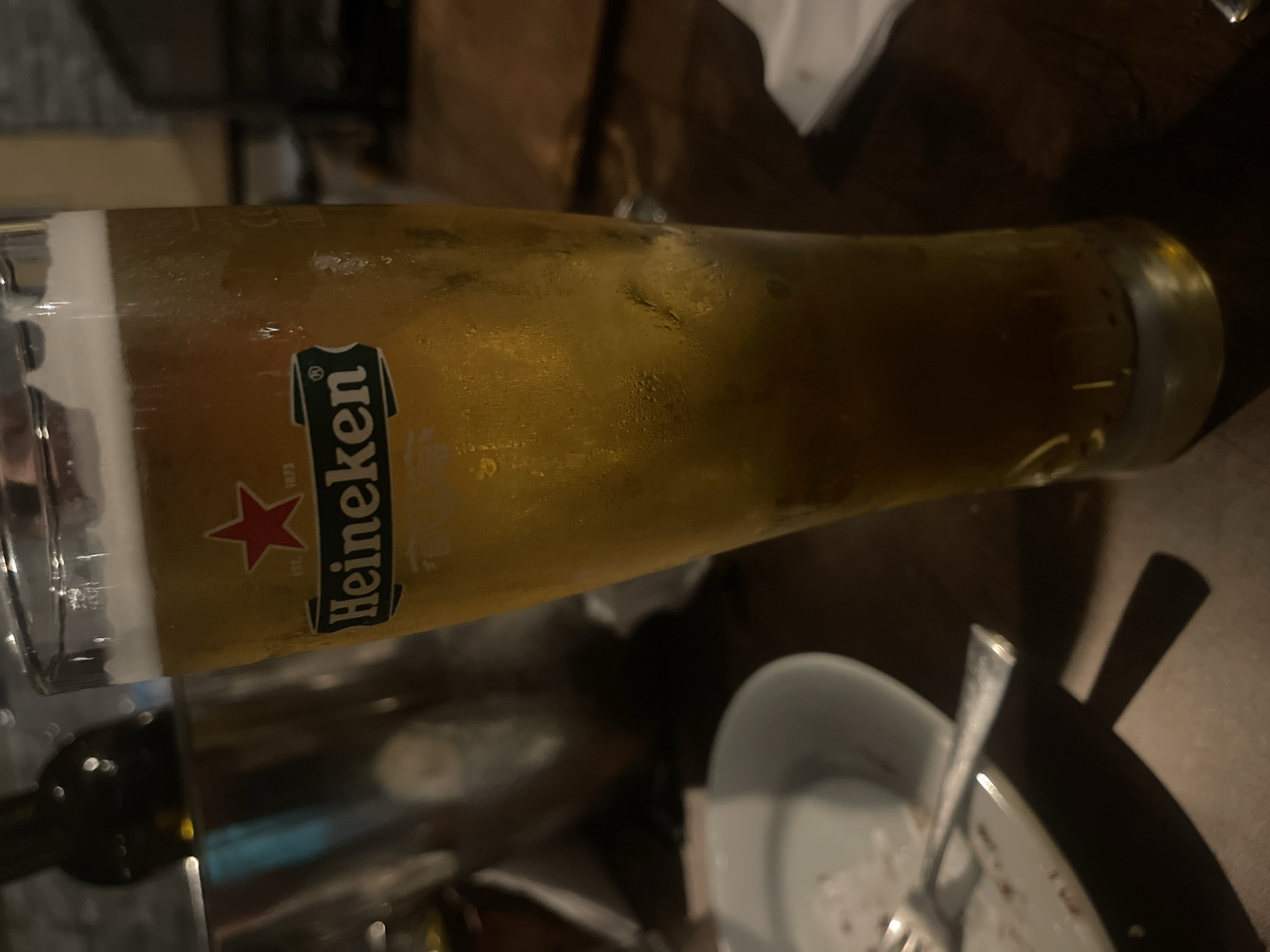 A close-up, slightly blurry, eye-level shot focuses on a tall, condensation-covered glass of Heineken beer. The beer is a light golden color with a frothy white head. The Heineken logo, a green rectangle with "Heineken" written in white letters and a red star above it, is clearly visible on the glass. The glass sits on a dark wooden table, with some of the condensation droplets reflecting the ambient light. To the left of the beer glass, a portion of a white plate with a fork resting on it is visible. In the background, out of focus, are a silver bucket, a dark bottle, and other dark, indistinct objects, suggesting a restaurant or bar setting. The lighting is dim, with warm tones, indicative of an indoor evening environment. The scene does not provide any information regarding the city of Ruse, Bulgaria.