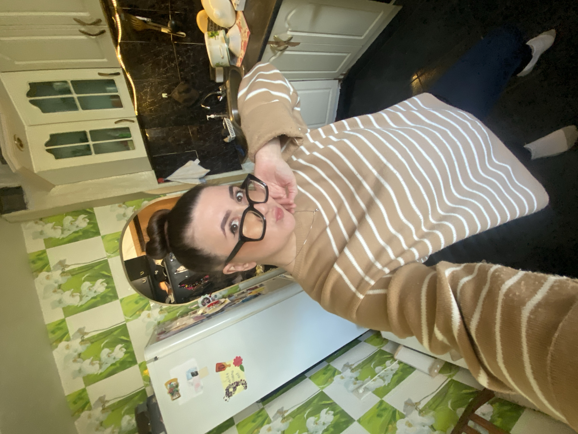 A woman with dark hair pulled into a bun and wearing large, black-rimmed glasses poses for a selfie in a kitchen. She is wearing a tan sweater with white horizontal stripes, dark blue pants, and white socks. She is standing in front of a white refrigerator adorned with magnets. Behind her and to the left is green and white checkered flooring, and a reflection of a kitchen counter and cabinets in a convex mirror. Above the refrigerator are off-white cabinets, and in the upper background, there are dark granite countertops. It is not possible to identify the specific location in Chaikovsky, Russia, from the image.