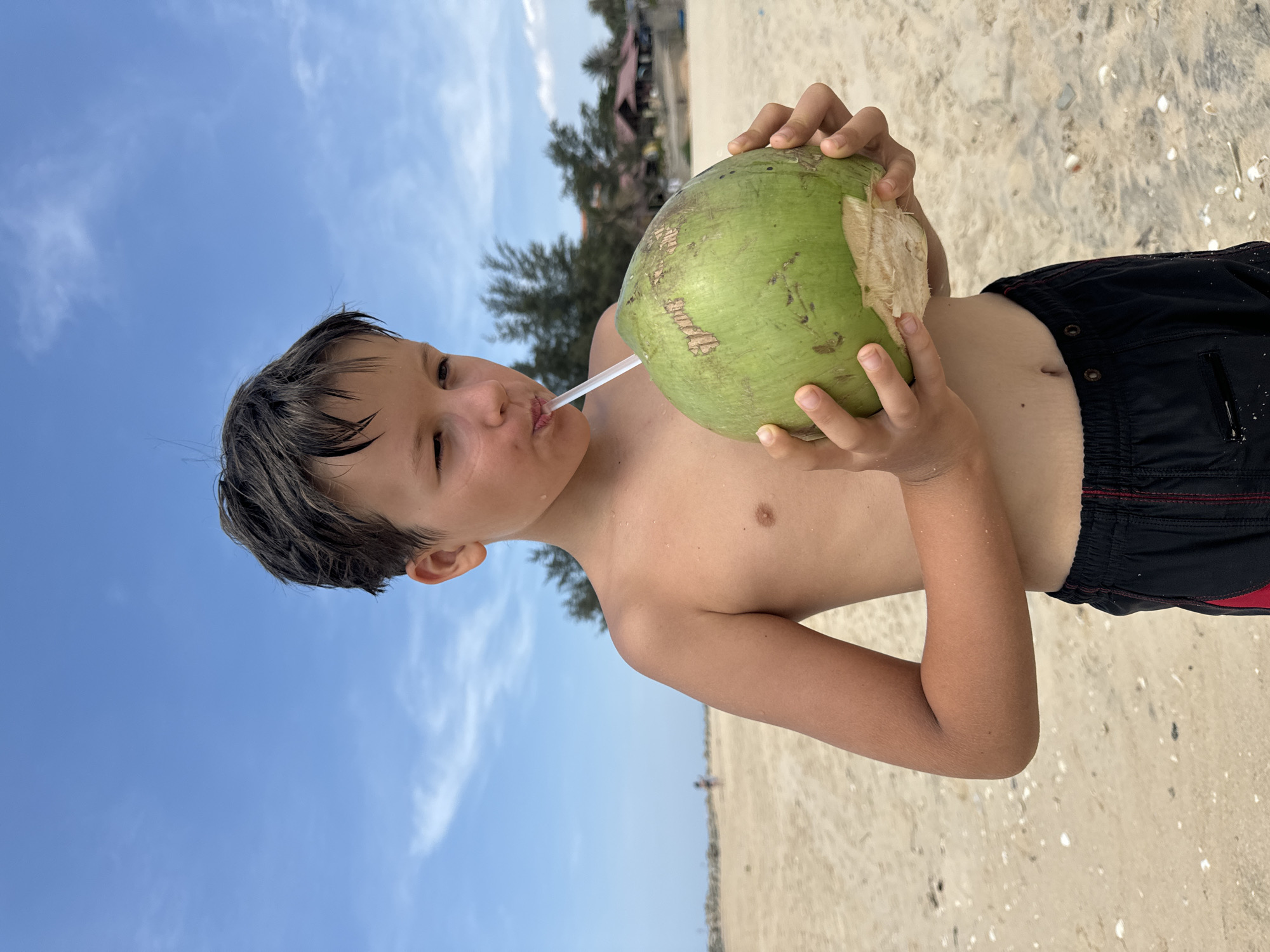 A child drinks from a green coconut with a straw on a beach. The child wears black swimming trunks. The beach is sandy, and there are trees and buildings visible in the background. The sky is blue with some clouds. The location is Phu Thuy Ward, Vietnam.