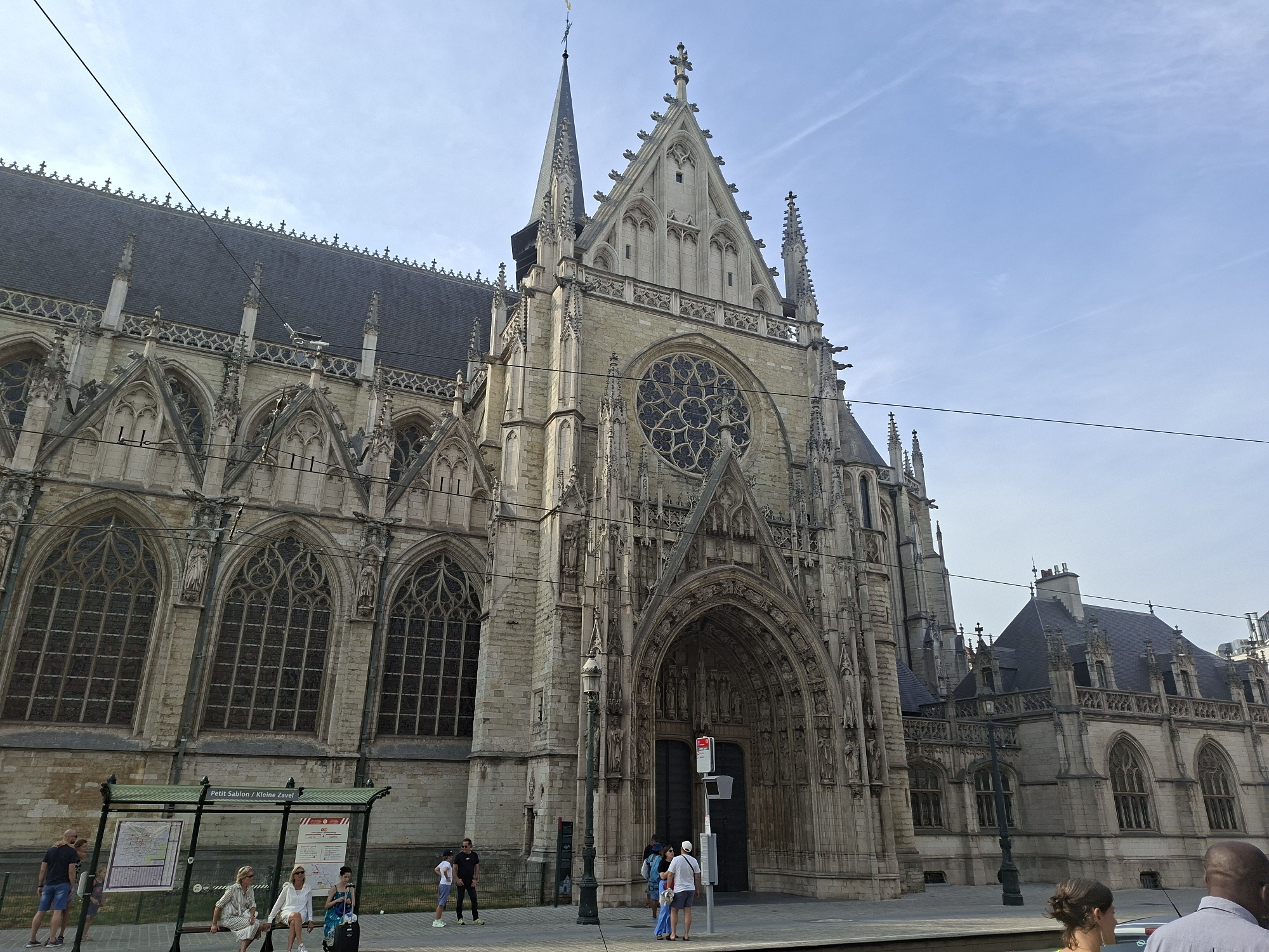 The image shows the exterior of the Saint-Jacques-sur-Coudenberg church, a prominent landmark in Brussels, Belgium. The church, constructed in a Gothic architectural style, features a large rose window above its main entrance and ornate carvings and spires. In the foreground, a bus stop sign with the inscription "Petit Sablon / Kleine Zavel" indicates the location is near the Place du Petit Sablon. Several individuals are visible, some waiting at the bus stop and others walking nearby. The sky is a clear blue with a few wispy clouds. The overall scene depicts a typical day in a European city, with a blend of historical architecture and modern urban life.