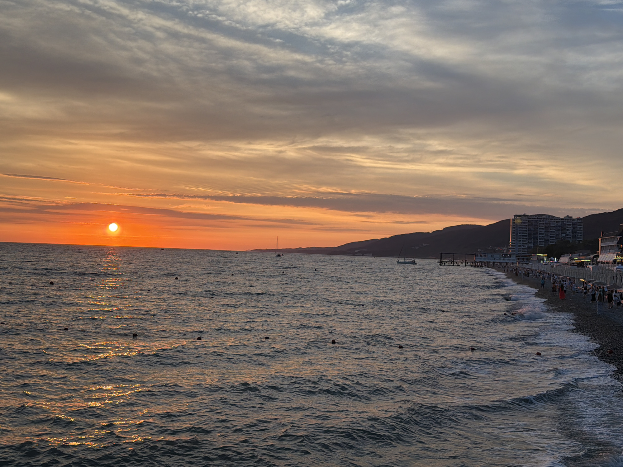 This is a wide-angle, eye-level, outdoor photograph capturing a sunset over the Black Sea near Sochi, Russia. The sun, a bright orange orb, is low on the horizon, casting a golden reflection across the rippling water. The sky is a canvas of soft orange, pink, and gray clouds, with some streaks of white.
In the middle ground, the coastline of Sochi is visible, characterized by a pebble beach lined with beach umbrellas and loungers. A few people are strolling along the shore, some appearing to be tourists. Several buildings, including a tall hotel, are situated along the coastline, nestled against the dark silhouettes of the mountains that rise behind the city. A pier extends into the sea, and a couple of sailboats are dotted on the water. Several buoys are visible in the sea, marking off swimming areas. The waves are small and gentle, lapping at the shore. The overall atmosphere is serene and picturesque, indicative of a pleasant evening on the coast.