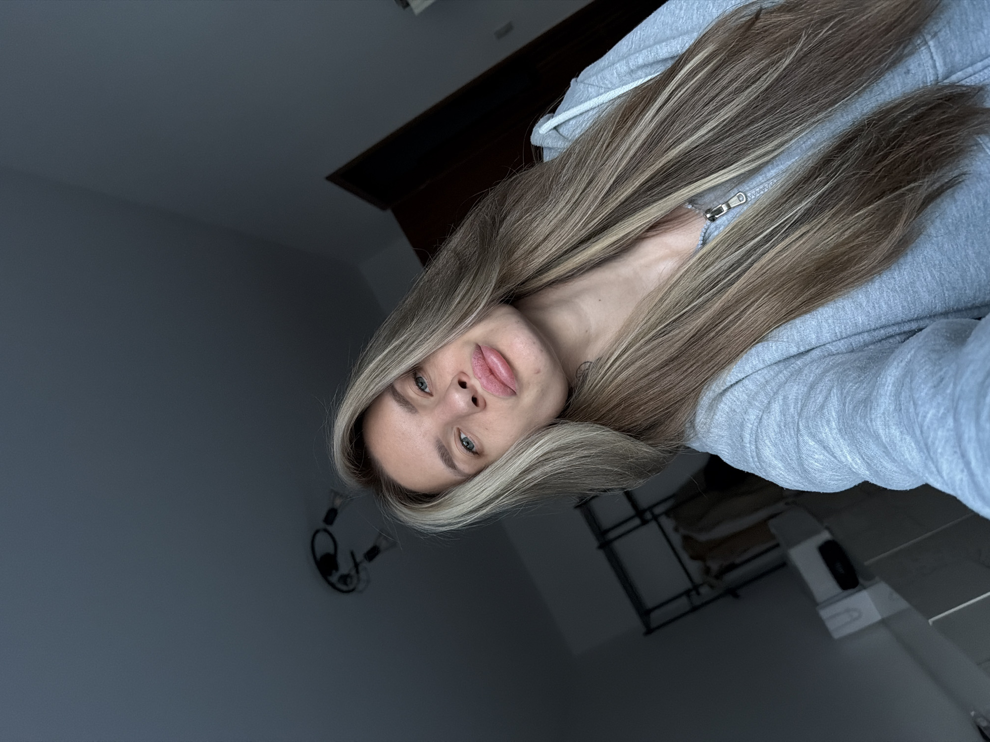 A young woman with long, highlighted blonde hair and blue eyes is pictured from a low angle, tilting her head to the right. She's wearing a light gray hooded sweatshirt, partially zipped. Behind her, a dark wooden headboard is visible above a white nightstand with an object resting on it. On the wall to the left hangs a black headphone. The room is dimly lit. The location cannot be determined as being in Berlin based on the provided image.