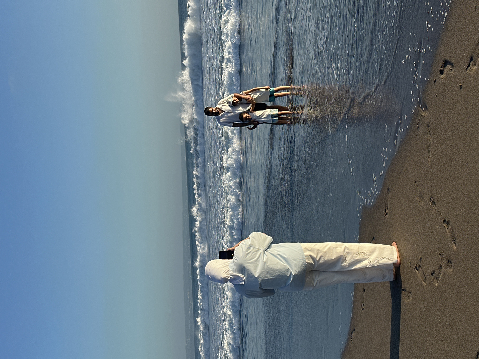This is a medium shot, eye-level, full-body photograph taken on a beach in North Kuta, Indonesia. The image captures a family of four posing for a photo in the shallow ocean water. A woman wearing a light blue, long-sleeved shirt and white, loose-fitting pants, along with a white hijab, stands on the sandy shore, facing the ocean. She is holding up a black smartphone in her right hand, appearing to take a photo.
In the water, a man and two young boys are standing. The man, wearing a white short-sleeved collared shirt and dark shorts, has his arm around one of the boys, who is wearing a white tank top and blue shorts. The other boy, dressed similarly in a white tank top and blue shorts, stands next to them. The boys are positioned in front of the man.
The ocean behind the family has moderate waves with white foam crests. The sky is clear and blue, with a slight haze visible on the horizon. The beach sand is dark brown and shows several footprints. The lighting suggests it is either early morning or late afternoon, with a warm, soft glow.