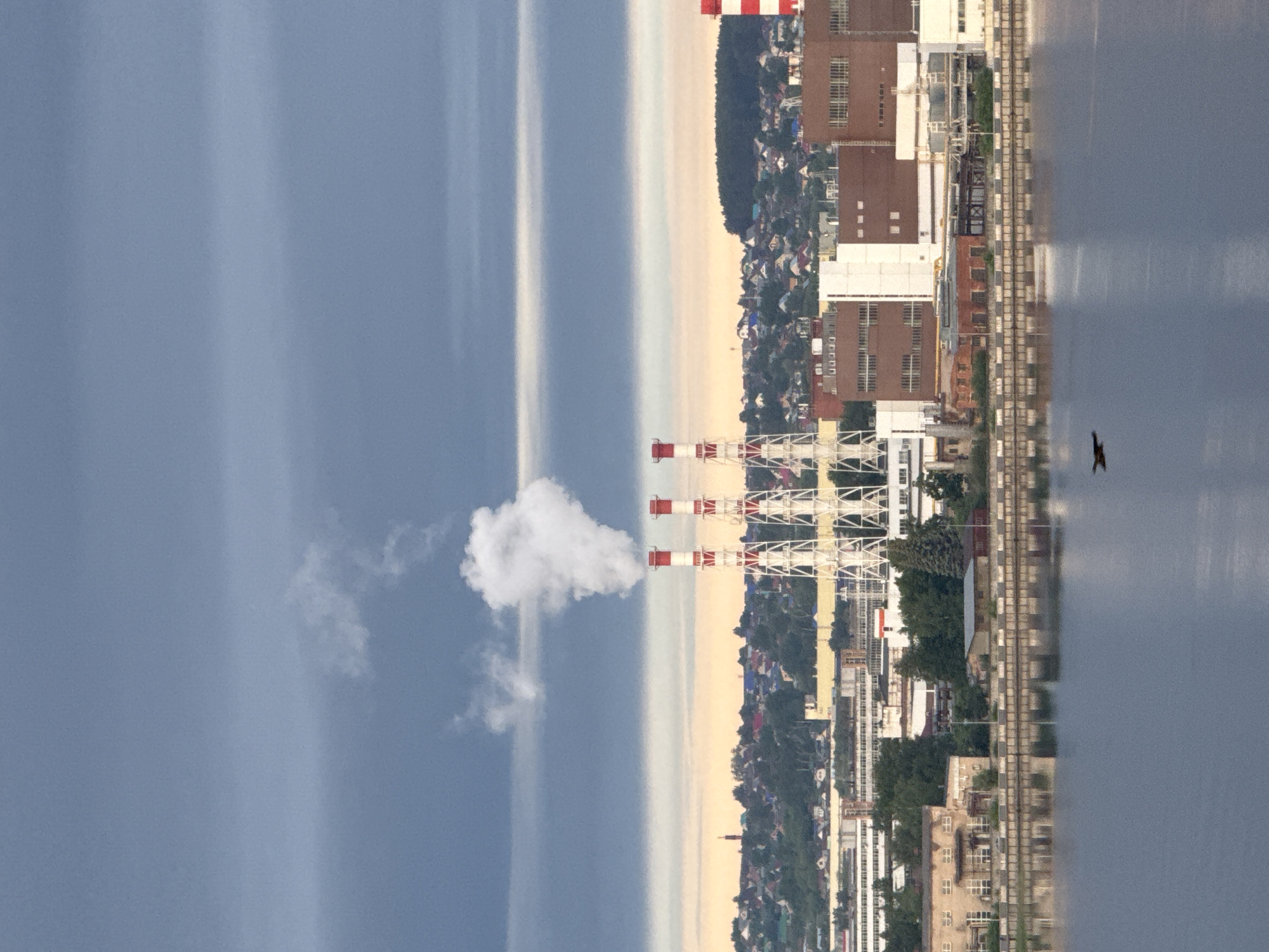 The image depicts an industrial scene in Izhevsk, Russia. Prominent are three red and white smokestacks emitting a plume of white smoke into a blue sky streaked with light. Adjacent to the smokestacks is a dense urban area with various buildings, some of which appear to be industrial. The edge of a river or large body of water is visible on the right side, with a bird flying above it. A railway runs along the waterfront.