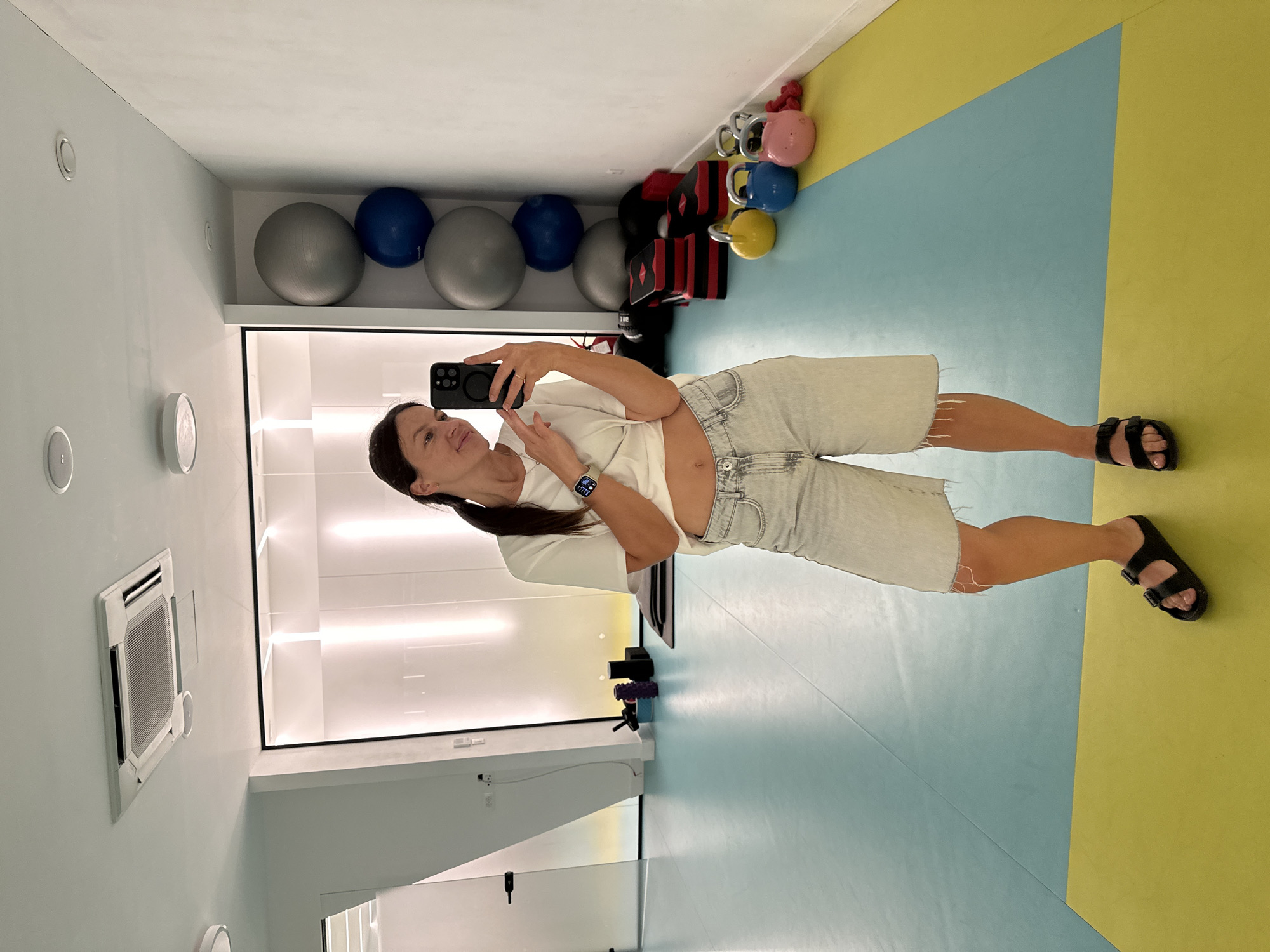 A woman with dark hair pulled into a ponytail, wearing a white t-shirt and light-wash denim shorts, takes a selfie in a gym. She is standing on a mat with a blue and yellow pattern. The gym appears to be equipped with fitness equipment, including large exercise balls, kettlebells of various colors, and what appear to be aerobic step platforms. There is an air conditioning unit visible on the ceiling and bright, vertical lighting in the background. The overall environment suggests a clean and modern fitness facility.