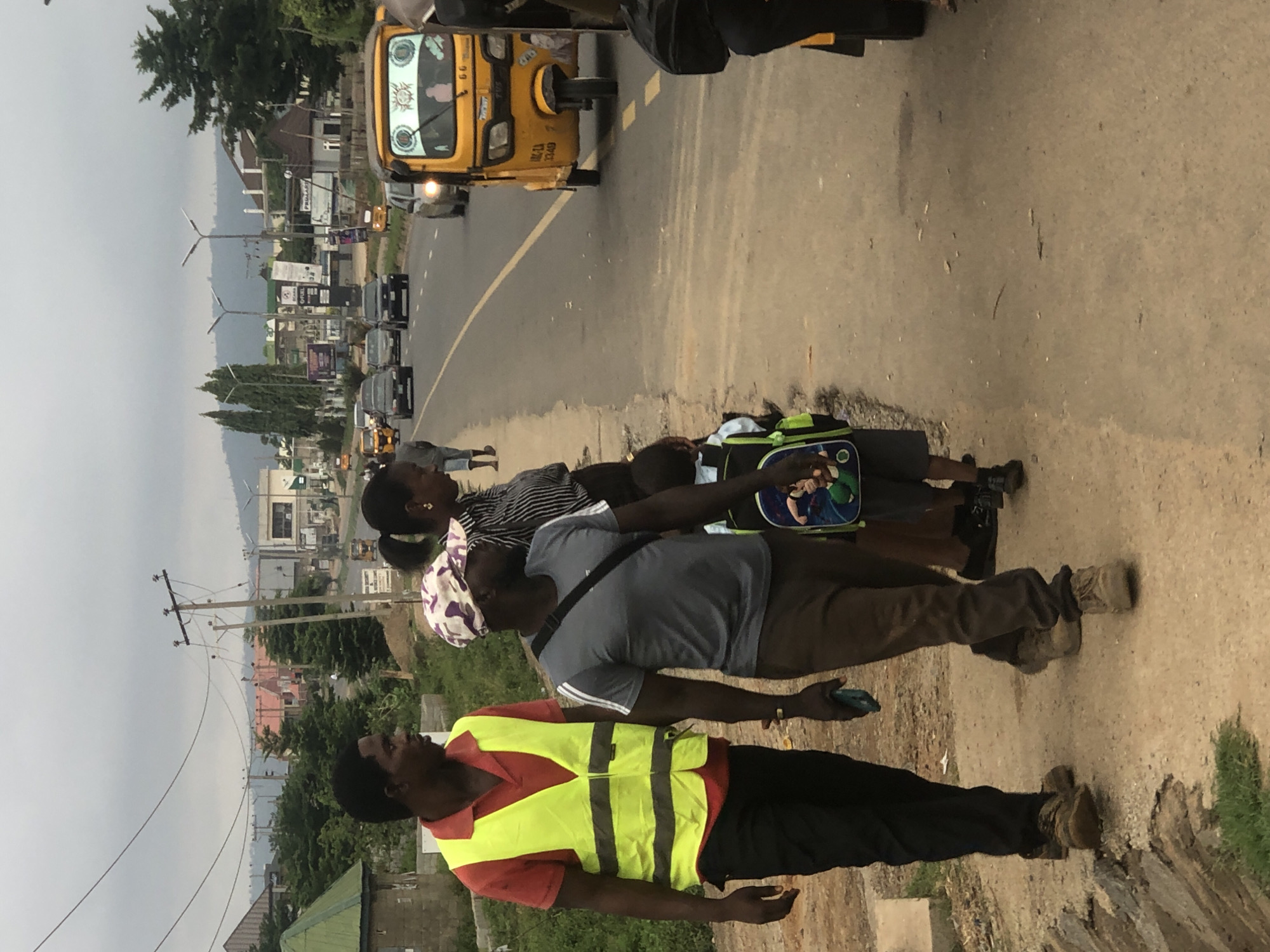 The image depicts a roadside scene in Abuja, Nigeria, with several individuals, a yellow tricycle (Keke), and vehicular traffic.
People: Four people are standing on a curb. The first is wearing a red shirt under a yellow reflective vest. The second wears a gray t-shirt with dark pants. The third wears a striped shirt and a head wrap. The fourth individual is wearing a backpack.
Objects: A backpack is visible on the ground next to the children. There are cars and yellow tricycles in the background.
Environment: The scene is along a roadside in an urban setting. Power lines and buildings are visible in the background. The sky appears overcast. The road is paved with a yellow divider line.