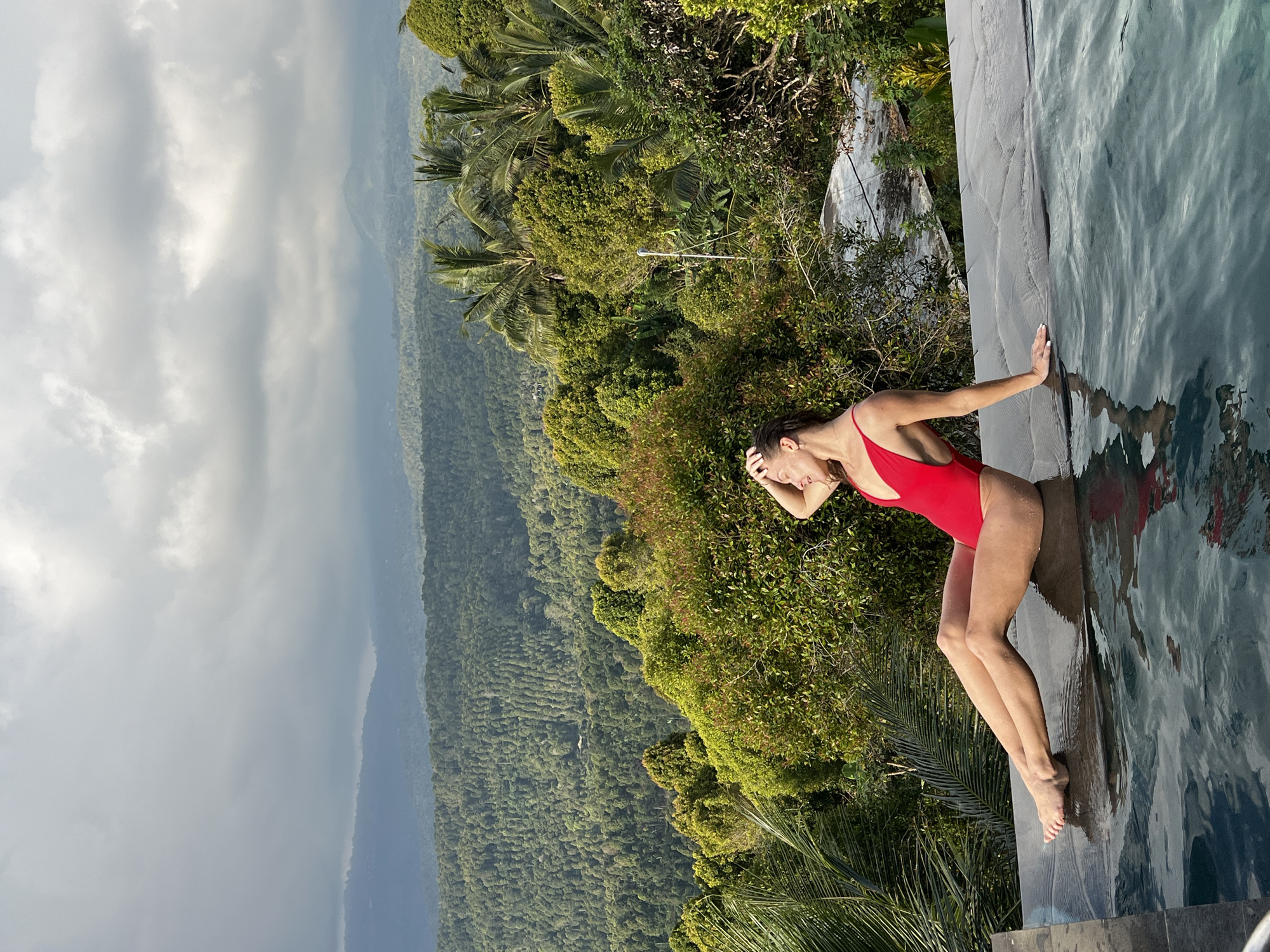 A woman wearing a red one-piece swimsuit is sitting on the edge of an infinity pool. Her legs are extended and her feet are in the water. She is looking to her left with her right hand touching her forehead. The pool overlooks a lush, green valley with rolling hills and dense forest. Palm trees and other tropical vegetation are visible in the foreground and throughout the landscape. The sky is overcast with dramatic clouds. The location is Busungbiu, Indonesia.