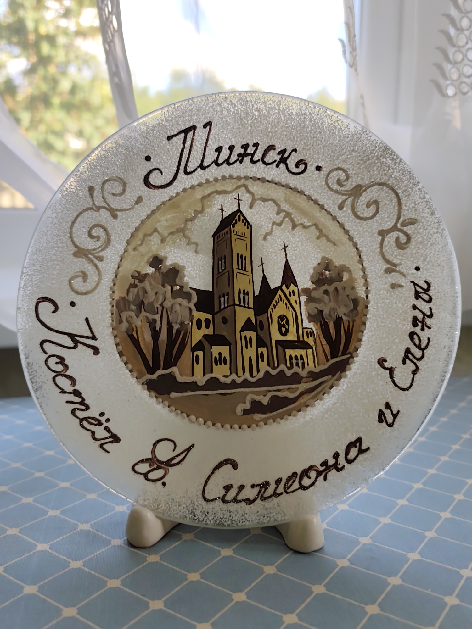 A round decorative plate with a painted image of a cathedral and text is displayed on a blue and white checkered tablecloth. The plate has a textured, frosted glass surface with a raised, glittery border. The central image depicts a stylized yellow and brown cathedral with multiple spires and windows, surrounded by trees. The text on the plate is in Cyrillic script and appears to be a dedication or inscription. The plate is supported by two small white feet. In the background, a white curtain with a decorative pattern is visible, with a blurred view of greenery and light shining through it, suggesting an outdoor setting behind the curtain. The plate is positioned on a surface that appears to be a table or shelf.