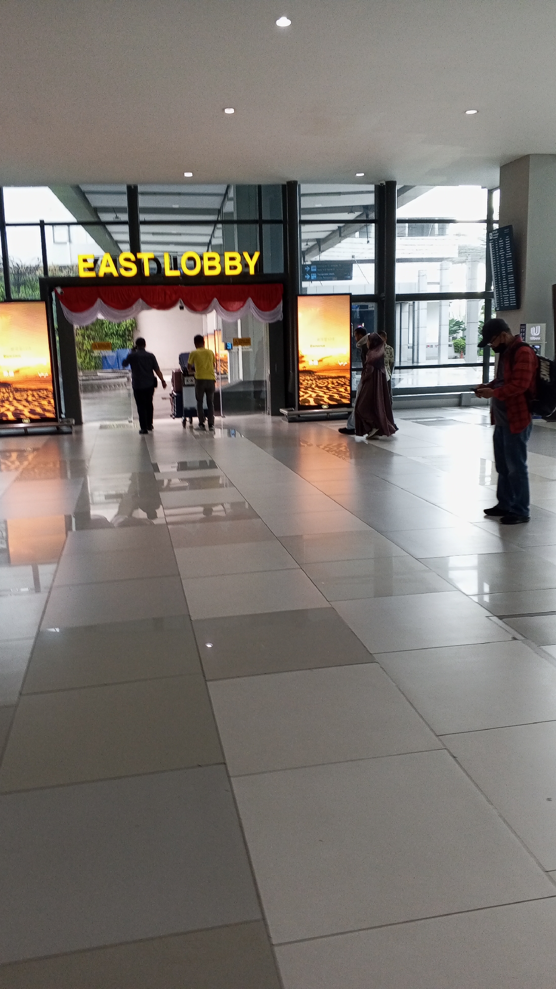 The image depicts the interior of what appears to be an airport or a large public building, specifically identified as the East Lobby of the city of Pajang, Indonesia. The scene is brightly lit by overhead lights and large digital screens displaying what seems to be scenic imagery. The floor is tiled with large, reflective square tiles in varying shades of gray and white.
In the foreground, the tiled floor stretches towards the background. In the mid-ground, near the entrance to the East Lobby, two individuals are seen walking away from the camera and into the lobby. One is wearing a dark shirt and pants, and the other, slightly ahead, is in a yellow shirt and gray pants, pushing a luggage cart.
Further to the right in the mid-ground, a person wearing a dark colored headscarf and a flowing robe is walking towards the right of the frame. To their right, a person wearing a plaid shirt over a darker shirt and jeans is standing, looking down at their phone and holding a backpack over their shoulder.
The East Lobby is clearly marked by large, illuminated yellow lettering above an arched entrance adorned with red and white decorative fabric. Large glass windows and doors are visible throughout the space, offering glimpses of the exterior. Signage indicating directions is also present on the right side of the image. The overall environment suggests a modern, clean, and functional public space.