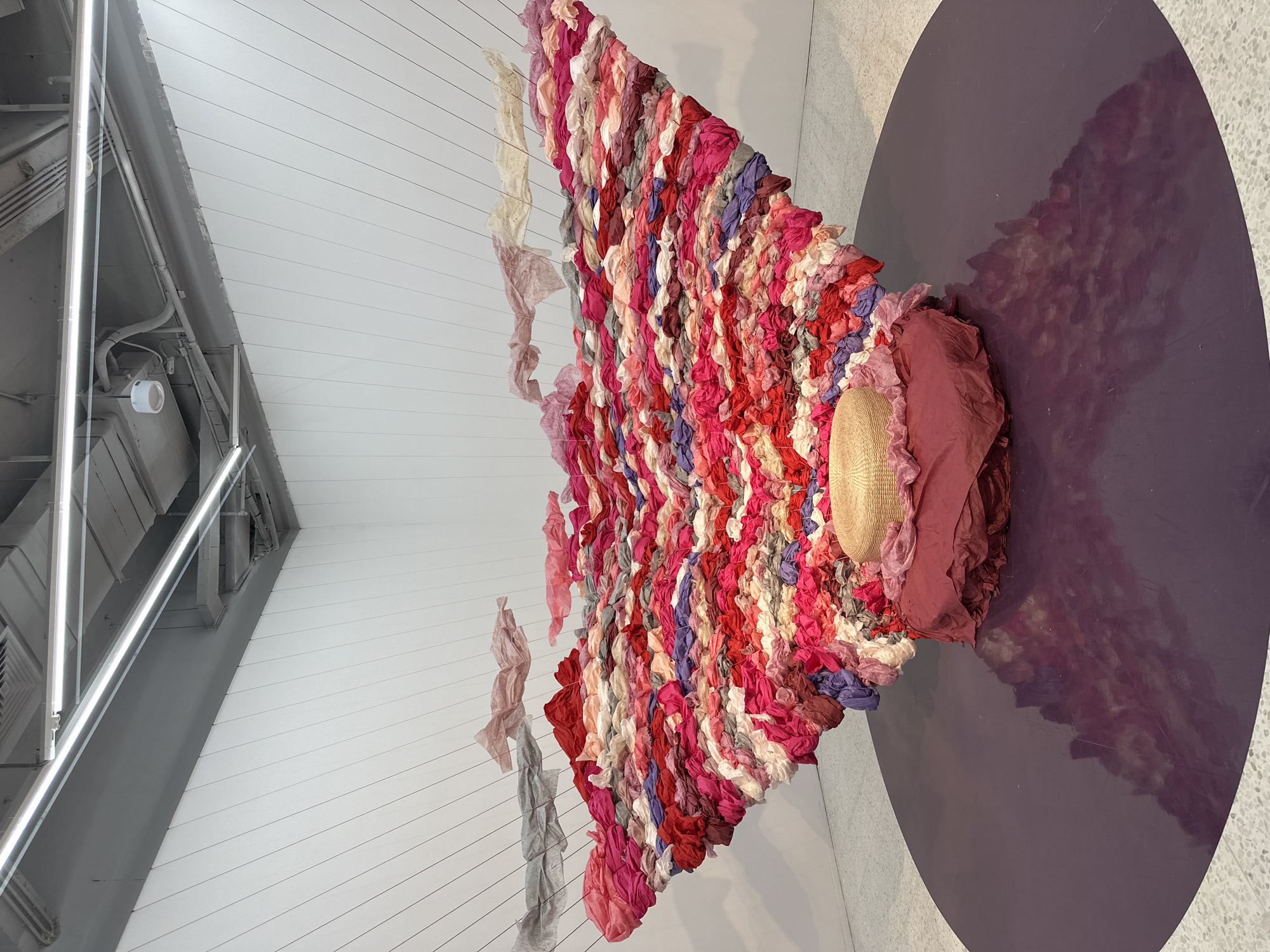 The image shows an art installation in an indoor space. The installation features a large, multi-colored textile arrangement composed of gathered fabric strips in shades of red, pink, purple, and white. These fabric strips are suspended from thin, horizontal lines attached to the ceiling. A round, woven hat sits amidst the fabric at one end, resting on a draped piece of reddish-purple fabric that spills onto a large, purple circular platform on the floor. The walls are white and the floor has a speckled pattern. There are fluorescent light fixtures attached to the ceiling.
It is not possible to identify the location as Banan, China from the information provided in the image.