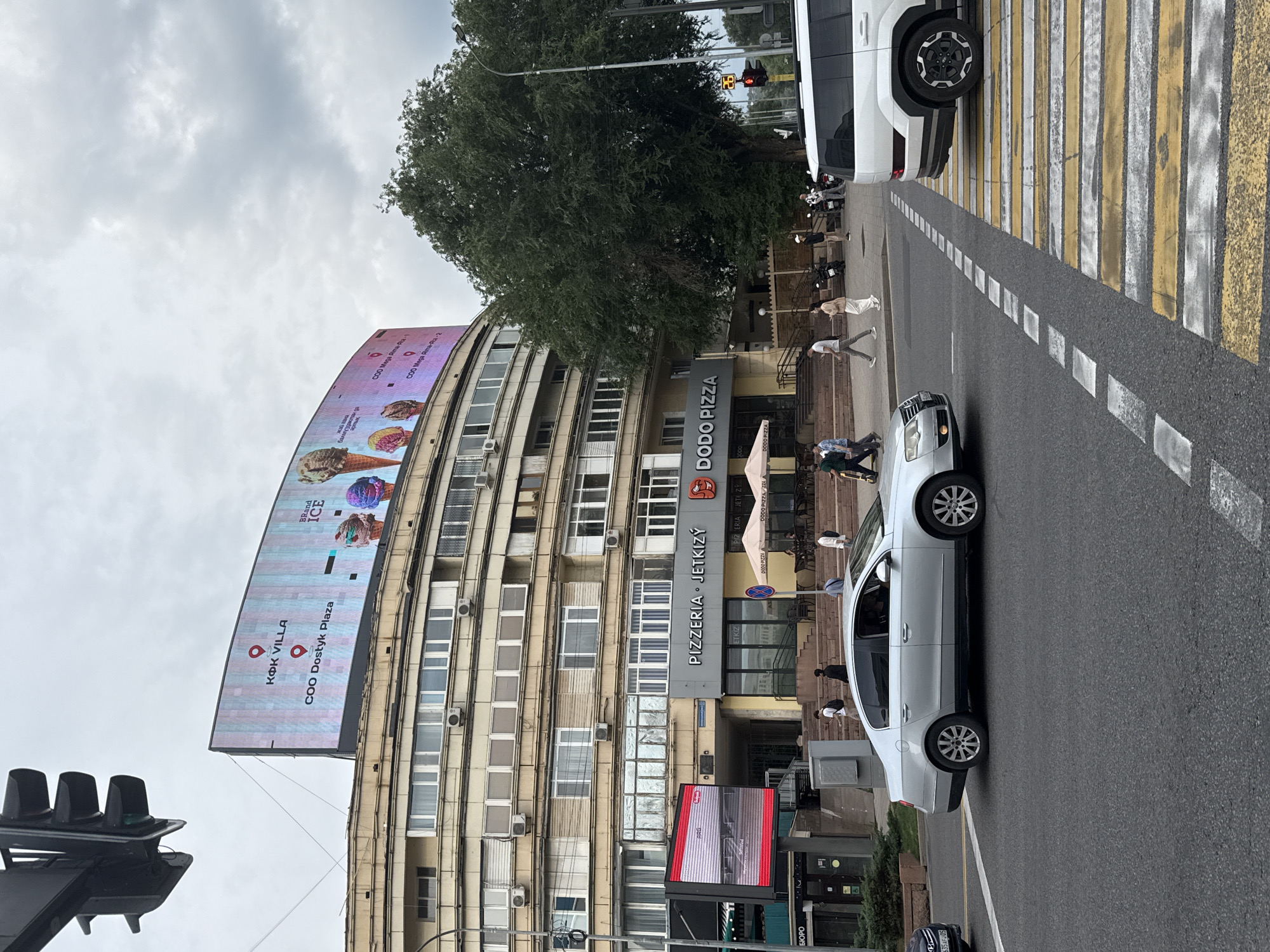 The image captures a street scene in Almaty, Kazakhstan. In the foreground, a silver Volkswagen Passat is turning at an intersection, with a white SUV waiting behind it at a crosswalk. The crosswalk is marked with yellow and white stripes. In the background, a large, curved building with multiple floors of windows and air conditioning units dominates the scene. A large digital billboard atop the building displays an advertisement featuring ice cream cones and text, including "KOK VILLA" and "COO Dostyk Plaza." Below the billboard, signage for "PIZZERIA JETKIZY" and "DODO PIZZA" is visible, along with several people walking on the sidewalk. A large, mature tree with dense foliage stands to the right of the building, partially obscuring the view of the sky. A traffic light with a red light illuminated is also visible to the right of the frame. The sky is overcast with clouds.