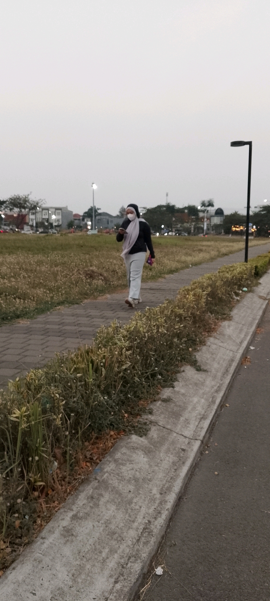 A full-body, medium shot captures a woman wearing a dark jacket, light-colored pants, and a light-colored hijab walking on a paved pathway. She also wears a white face mask. She is holding a dark object, possibly a phone, in her right hand, and a pink item in her left hand. The woman is walking away from the viewer on the right side of the frame, on a pathway that runs parallel to a road. The pathway is bordered on the left by a grassy field with some dry vegetation and on the right by a curb and a road. In the background, there are buildings, trees, and streetlights. The sky is overcast and appears to be dusk or dawn. The scene is likely in the city of Gunung Putri, Indonesia.