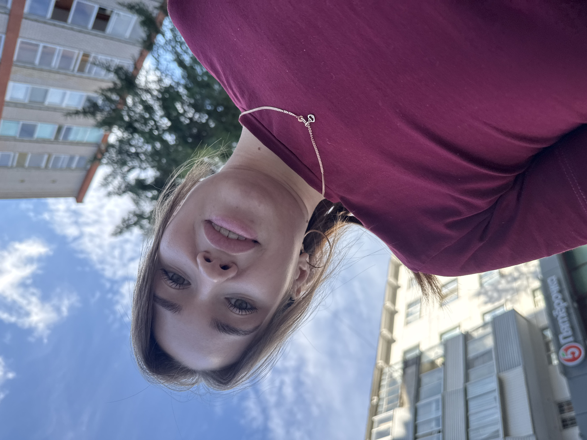This is a selfie taken outdoors, presumably in Chaikovsky, Russia. The subject is a young woman with light brown hair, looking directly at the camera. She is wearing a dark pink long-sleeved shirt and a delicate chain necklace with a small heart pendant. Her eyes are open, and she has a subtle smile. She is wearing hoop earrings.
In the background, there are buildings and trees. One building appears to be an apartment complex, with multiple windows visible. Another building, to the right, has a prominent sign with a red circle containing a white "5" and the Russian word "Пятёрочка" (Pyaterochka), which is a common Russian supermarket chain. The sky is blue with some white clouds, suggesting it is daytime and the weather is fair. The selfie is taken from a low angle, with the sky and buildings appearing above the woman's face, giving a sense of looking up. The overall mood is casual and perhaps a bit playful, given the selfie format.