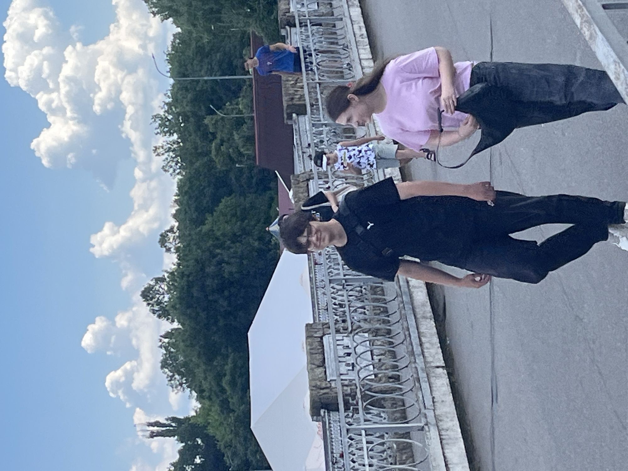 The image shows a group of people walking on a bridge. In the foreground, a young man in a black t-shirt and pants is walking towards the camera, with a black crossbody bag. To his right, a young woman in a pink t-shirt and dark pants is walking away from the camera, holding a black handbag. Behind them, other people are visible walking on the bridge, including a child in a patterned tank top and shorts, and a man in a blue t-shirt. The bridge has a metal railing with decorative elements and stone accents. In the background, there is a large white tent structure and trees. The sky is blue with fluffy white clouds. The scene appears to be a public outdoor space, likely a park or waterfront area. The presence of a white tent suggests a temporary event or market. The architecture of the bridge and the style of the buildings in the background could indicate a location in Krasnodar, Russia, as the lighting and vegetation are consistent with a temperate climate.