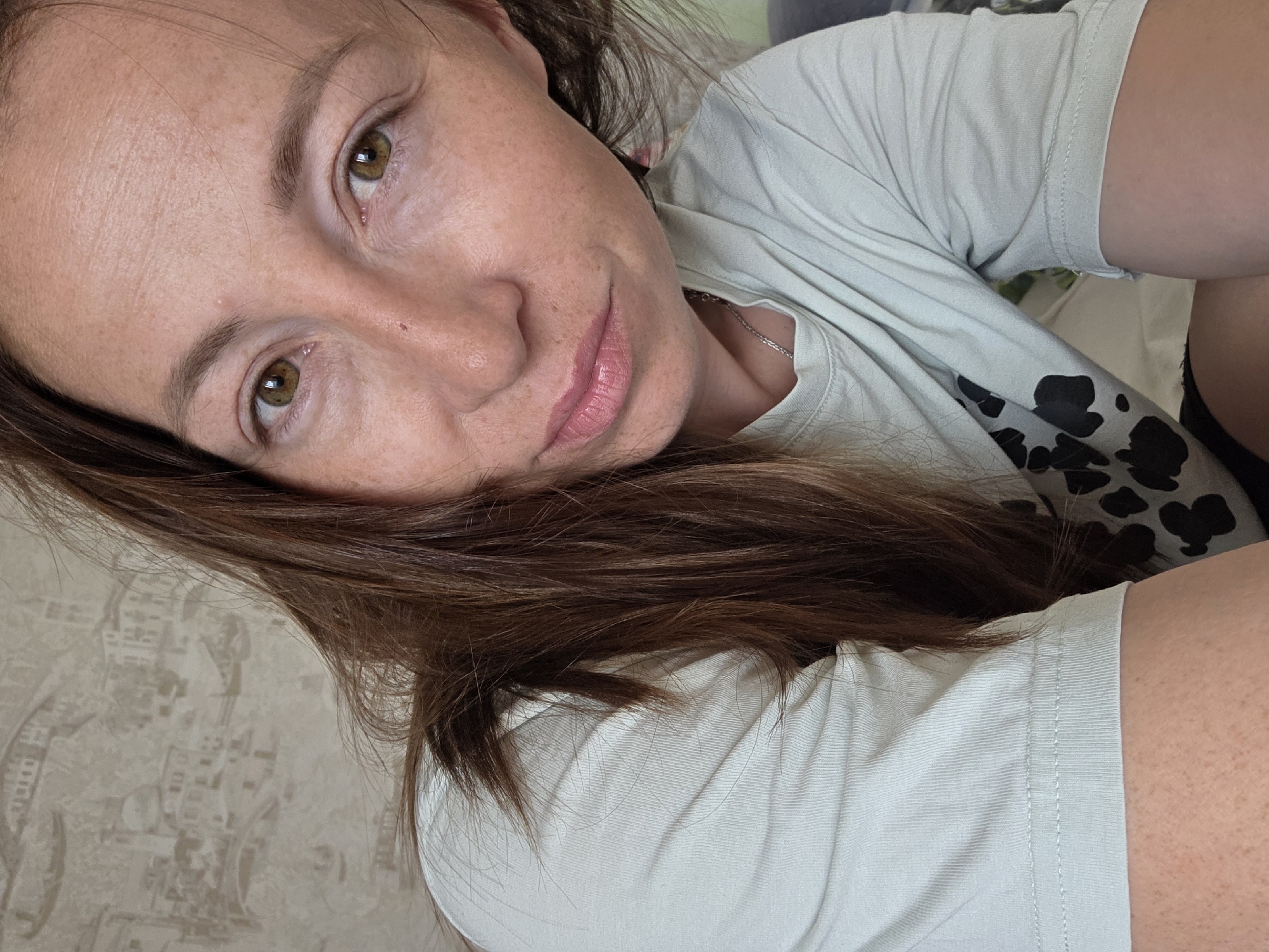 The image is a close-up selfie of a woman with brown hair and green eyes. She is wearing a light blue t-shirt with a black and white pattern on it. Her hair is long and falls over her shoulder. The background is a wall with a textured pattern that resembles buildings and arches. The lighting is natural and soft. The woman is looking directly at the camera with a neutral expression. There is no discernible location in Chaikovsky, Russia, or any other specific location.