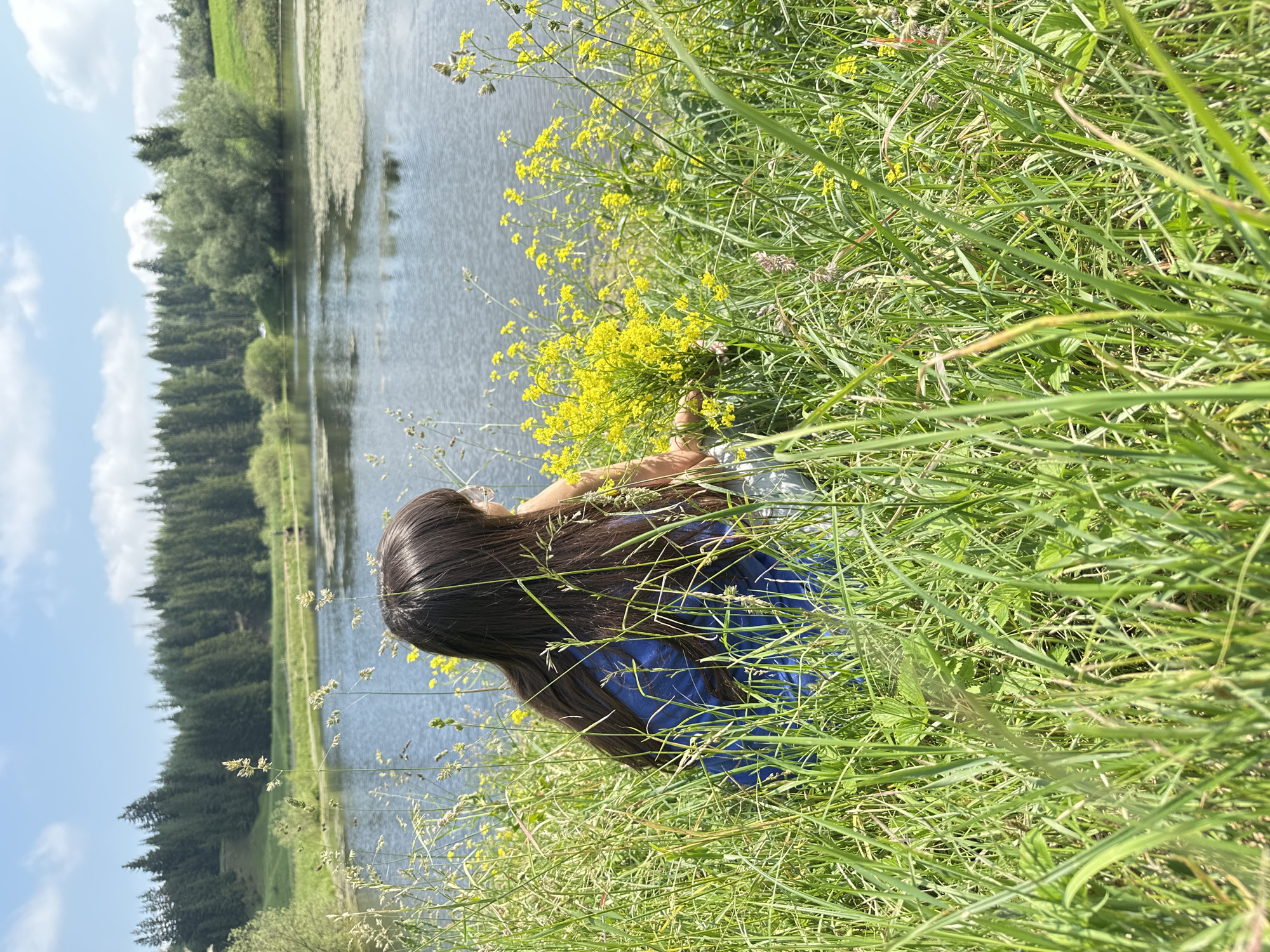 The image captures a serene natural scene with a person sitting in tall grass near a body of water. The person, with long dark hair, is wearing a blue top and is facing away from the camera, looking out towards the water. They are surrounded by lush green grass and yellow wildflowers, suggesting a springtime or summer setting. In the background, a calm lake is visible, reflecting the blue sky dotted with white clouds. Beyond the lake, a dense forest of evergreen trees lines the horizon, with rolling green hills on the left. The overall impression is one of peaceful contemplation in a natural landscape. The image does not contain any identifiable landmarks or context to place it specifically within Buranovo, Russia.