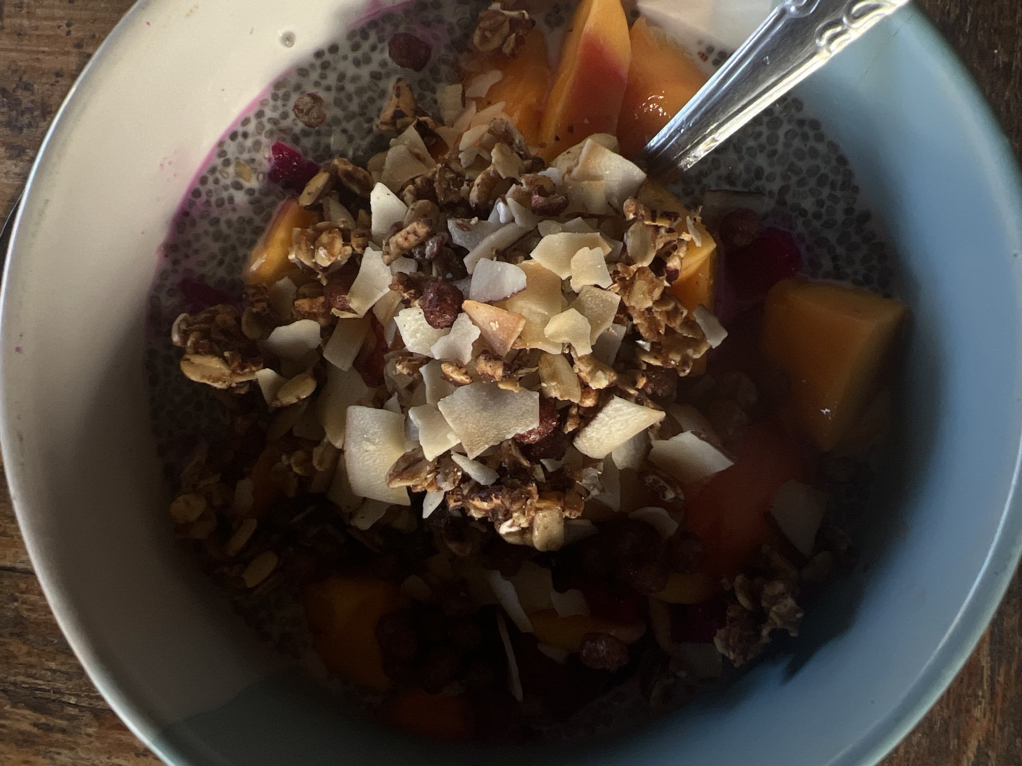 An overhead, close-up shot captures a vibrant and healthy breakfast bowl, possibly a chia seed pudding, placed on a wooden surface. The main subject is a light blue bowl filled with various ingredients.
The base of the dish appears to be a chia seed pudding, visible as a greyish, speckled mass, particularly around the edges of the bowl and beneath the toppings. Swirls of bright pink or purple, likely from dragon fruit, are mixed into the chia pudding on the left side.
Dominating the center of the bowl is a generous topping of crunchy granola, characterized by clusters of oats, nuts, and possibly dried fruit pieces, in shades of golden brown and dark brown. Mixed within and on top of the granola are numerous white, thin flakes of toasted coconut, adding texture and visual contrast.
Scattered throughout the bowl and partially submerged in the chia pudding are bright orange chunks and slices of fresh mango, indicating a tropical influence. Smaller, deep red or purple pieces of dragon fruit are also visible, complementing the larger fruit pieces.
A silver spoon, with an ornate handle design, is partially visible on the right side of the bowl, resting in the food, suggesting the meal is ready to be eaten or is already being consumed. The lighting is natural, highlighting the textures and colors of the food. No text is visible in the image. Given the ingredients, the meal suggests a breakfast or healthy snack, consistent with a tropical location like North Kuta, Indonesia.
