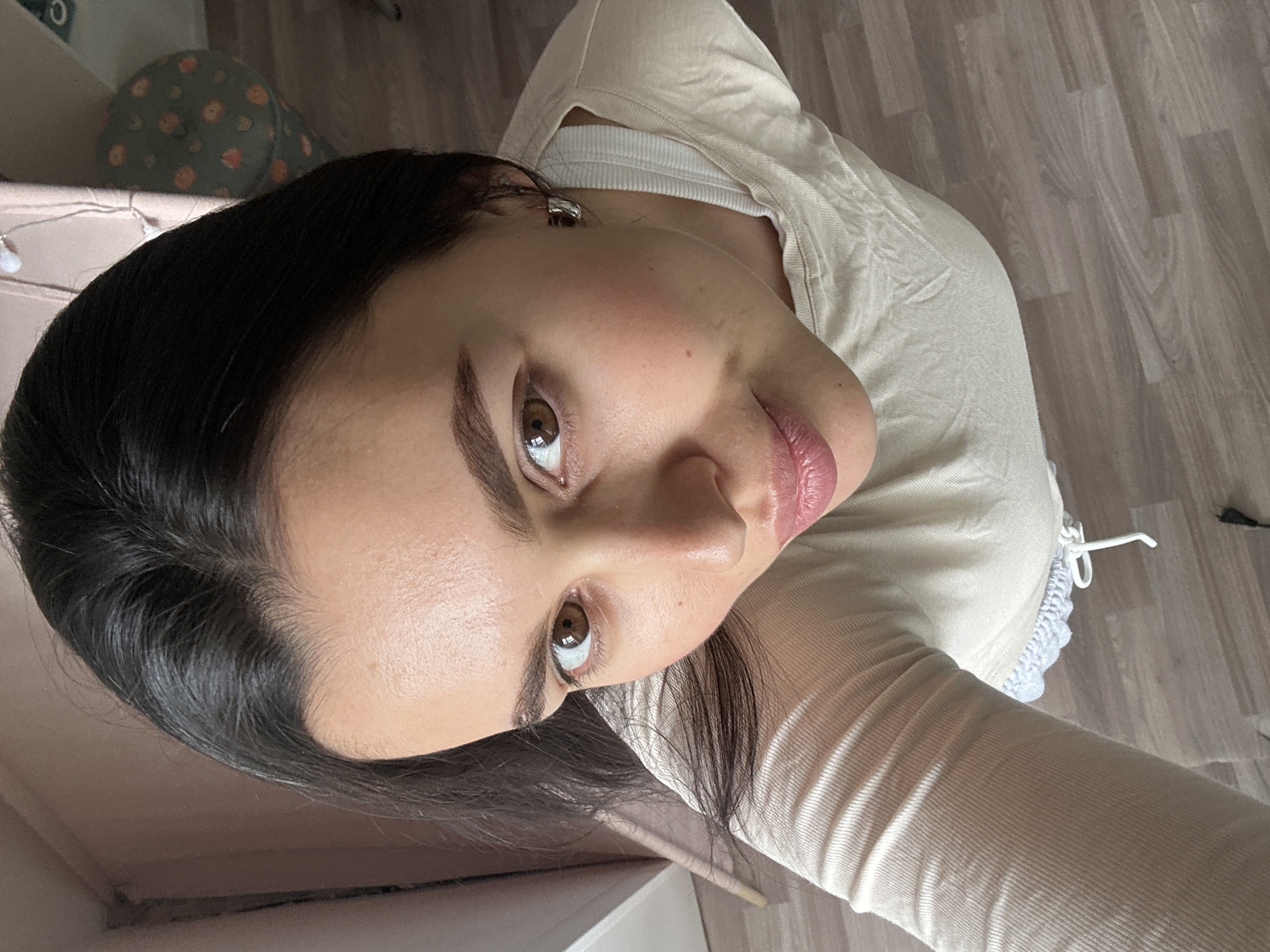 A woman is seen from an upward angle, possibly a selfie. She has dark hair, brown eyes, and fair skin. She wears a light-colored, ribbed shirt. She seems to be indoors, standing on what looks like laminate flooring. A piece of furniture and a cushion are visible in the periphery. The photo was likely taken in a domestic setting but there's no indication of location within Chaikovsky, Russia.