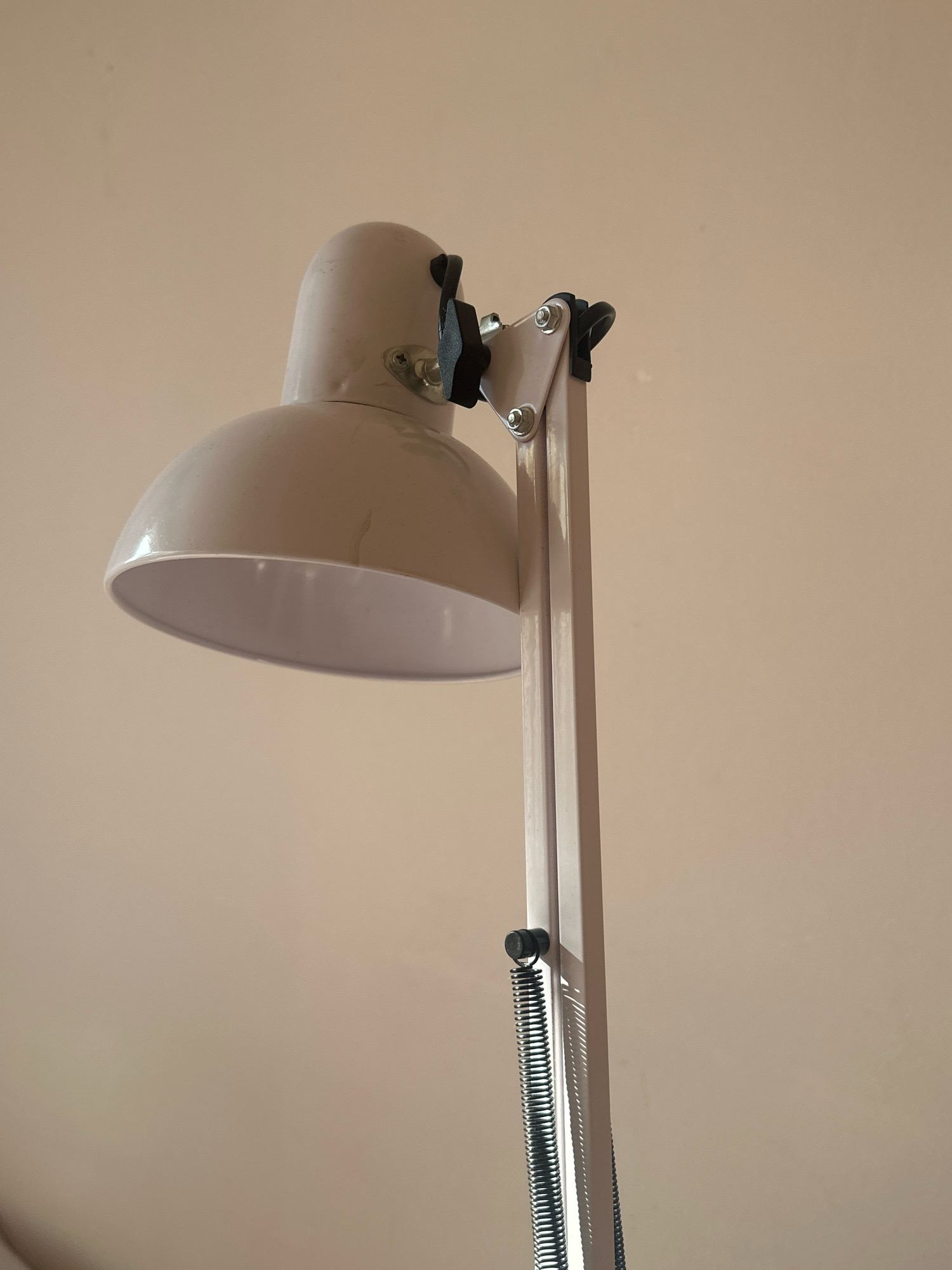 The image shows a close-up, slightly angled view of a cream-colored desk lamp against a plain, light brown wall. The lamp has a bell-shaped shade and a multi-jointed arm with visible springs. The background is uniformly lit and shows no discernible features, suggesting an indoor setting. There are no people or discernible actions captured in the image. The scene appears to be an interior residential or office space, but its specific location within the city of Gyumri, Armenia, cannot be determined from the visual information provided.