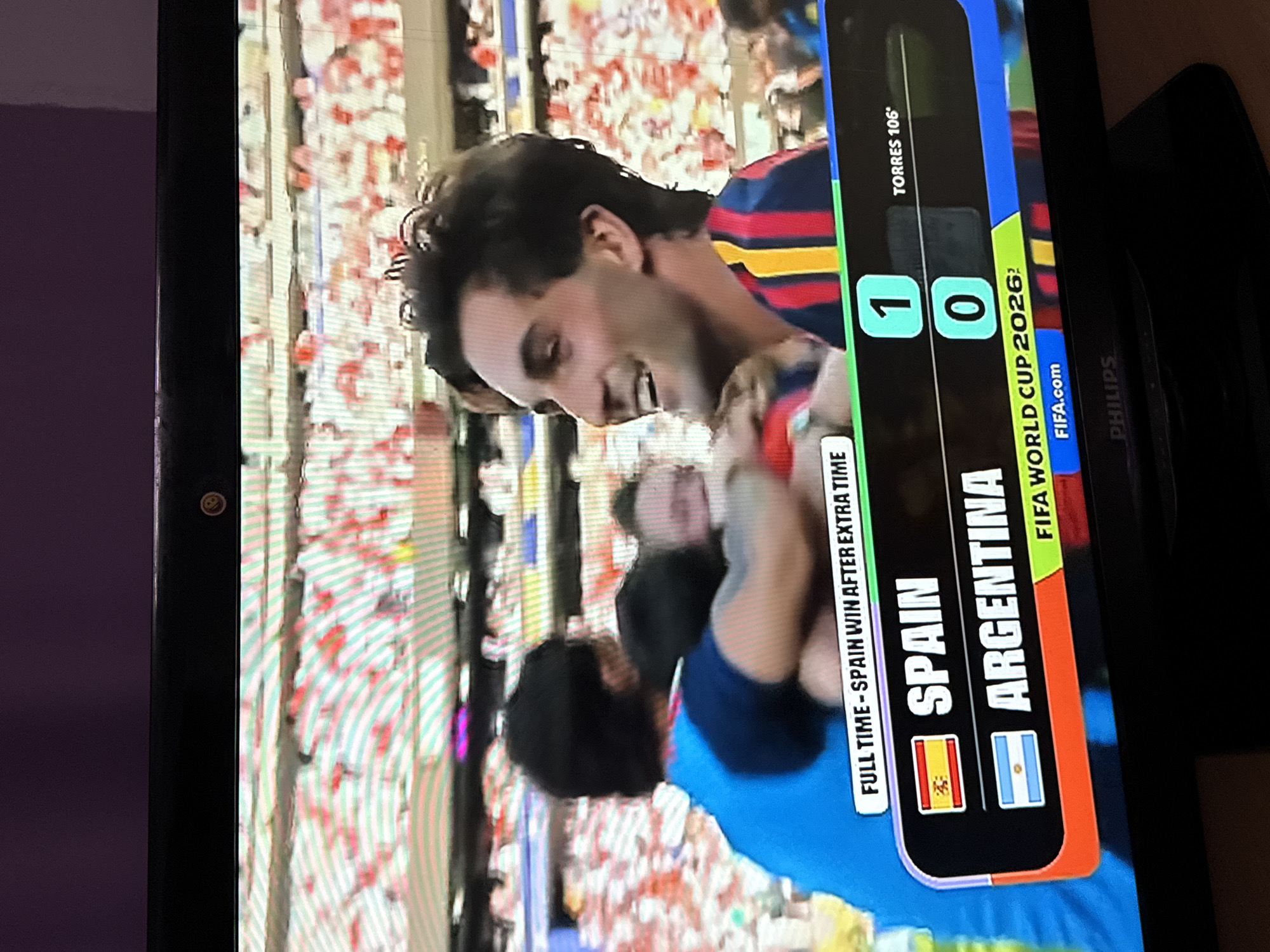 The image shows a television screen displaying a football match score. The screen shows that Spain won against Argentina with a score of 1-0, after extra time. The match appears to be part of the FIFA World Cup 2026. In the foreground, three men in football jerseys are celebrating, embracing each other. One man, in the center and facing the camera, is smiling broadly. The background shows a stadium filled with spectators. The television is placed on a stand in a room with a purple wall in the background and a Philips logo visible on the bottom of the stand. The specific location of Ufa, Russia, is not indicated in the image.