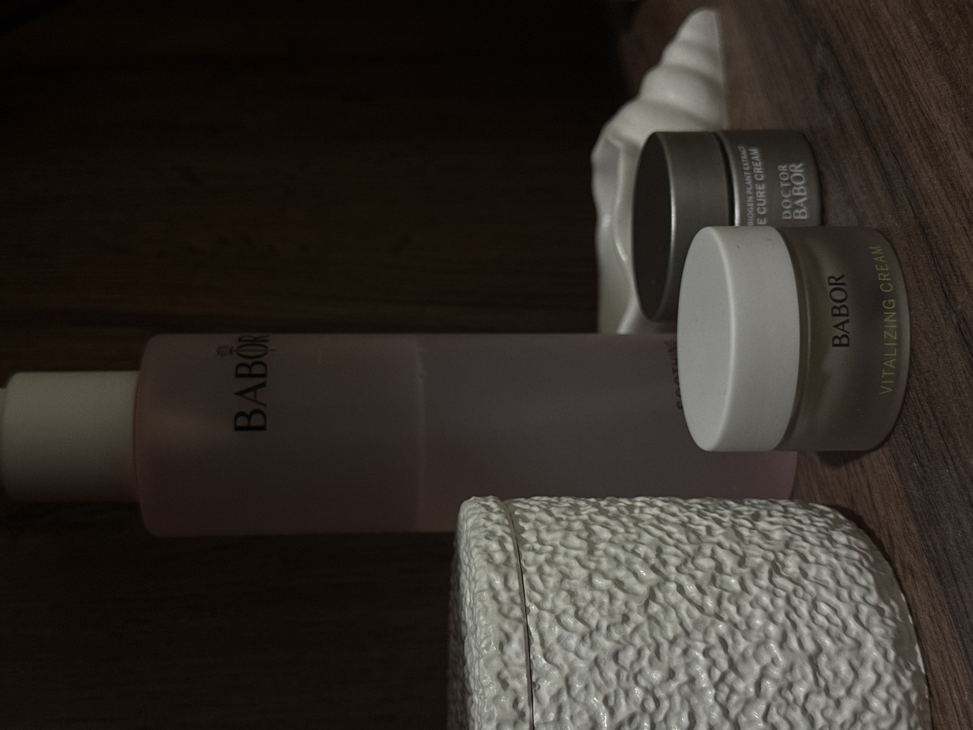 This close-up, dimly lit image captures an arrangement of skincare products on a dark brown wooden surface, possibly inside a cabinet or on a shelf, and is oriented as if rotated 90 degrees clockwise.
In the center-left, a tall, translucent pink bottle with a white pump cap is visible, featuring the brand name "BABOR" prominently. To its right, a smaller, round jar with a white lid and a translucent yellowish-brown base is positioned, labeled "BABOR VITALIZING CREAM". Behind this, another dark grey or metallic jar with a black lid is seen, bearing the text "BIOGEN PLANT EXTRACT E CURE CREAM" and "DOCTOR BABOR".
A white, textured object with a ridged or bumpy surface is partially visible in the bottom right corner, while a white, wavy-edged object is partially seen behind the metallic jar in the top right. The wooden surface displays a distinct grain. The overall low light suggests an indoor setting with poor illumination. No activity is taking place. The external context indicates the location is Chaikovsky, Russia, though this is not visually apparent from the image itself.