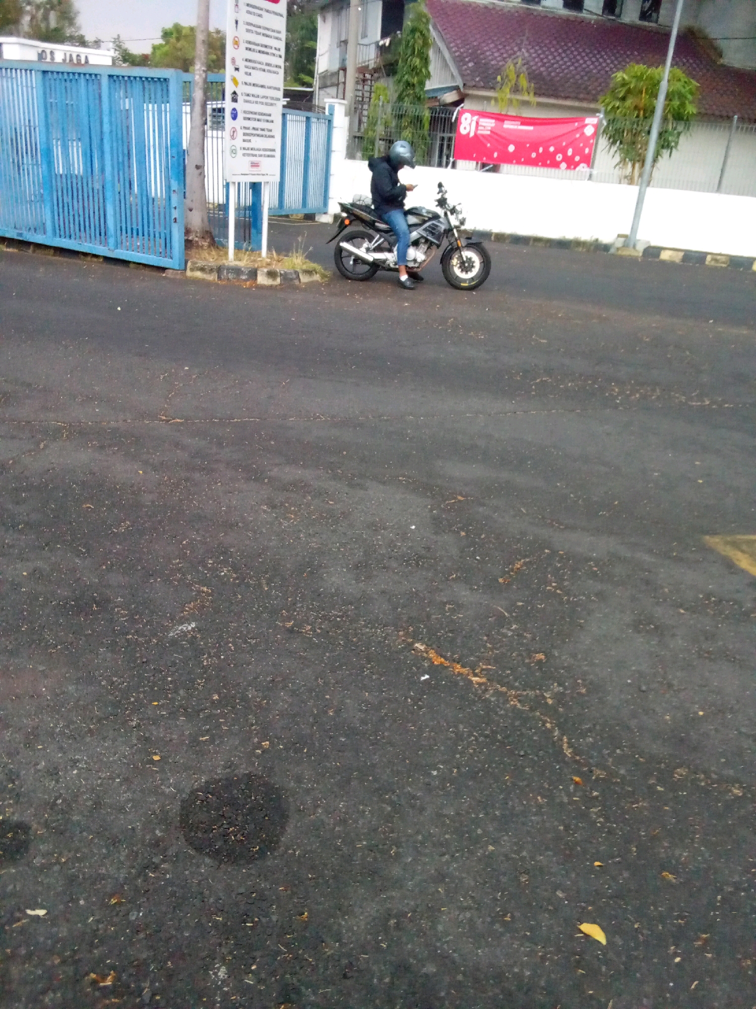 A person is sitting on a dark-colored motorcycle parked on an asphalt road in Mangli, Indonesia. The person is wearing a blue helmet, a dark hoodie, and blue jeans. They are holding a smartphone in their left hand. The motorcycle has a silver fuel tank and black bodywork. In the background, there is a blue metal gate with vertical bars, a white wall, and a building with a red-tiled roof. A red banner with white polka dots and the number "81" is hanging on the wall. Several trees and a signpost are also visible. The ground is a dark asphalt surface with some scattered leaves and debris.