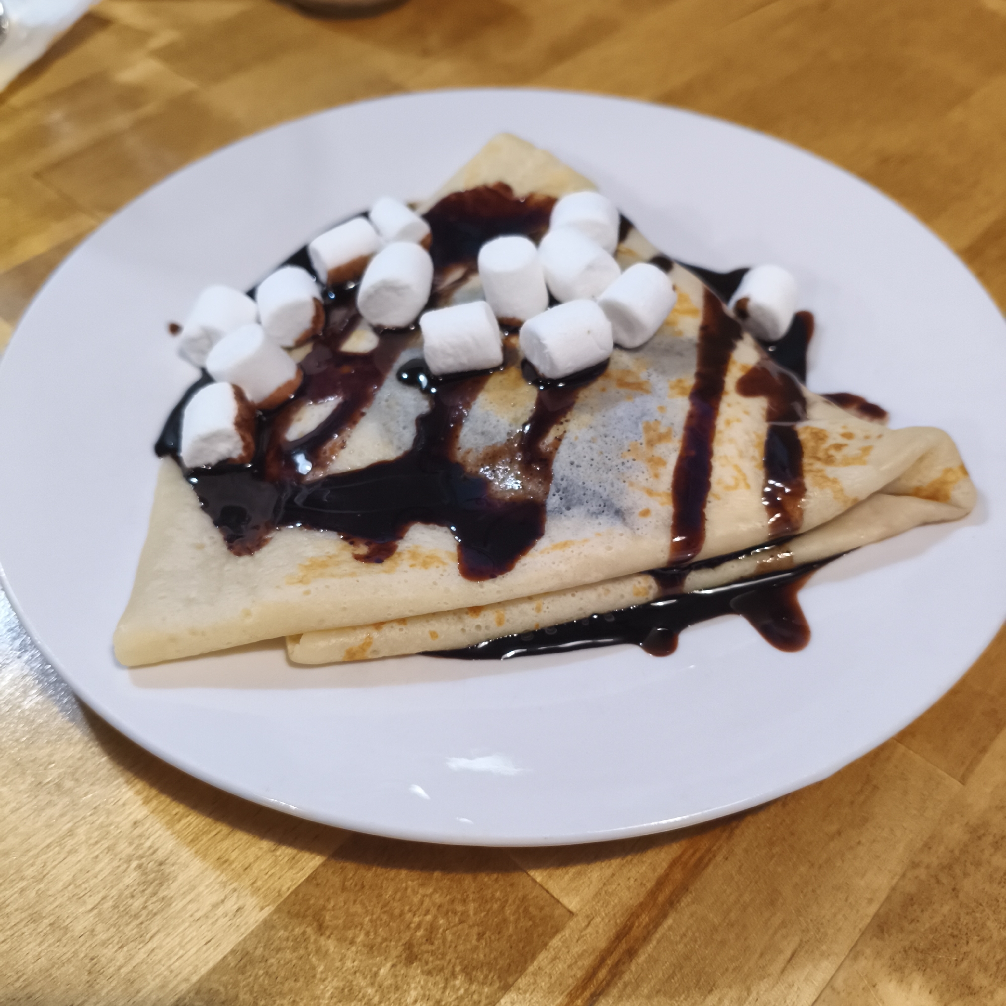 A triangular crepe, folded twice, rests on a white plate on a wooden table. The crepe is drizzled with chocolate sauce and topped with white marshmallows. The location is unknown. There is no information about people, subjects, actions, or interactions. The environment consists of the crepe, chocolate sauce, marshmallows, plate, and table. There is no evidence to suggest that the image was taken in the city of Chaikovsky, Russia.