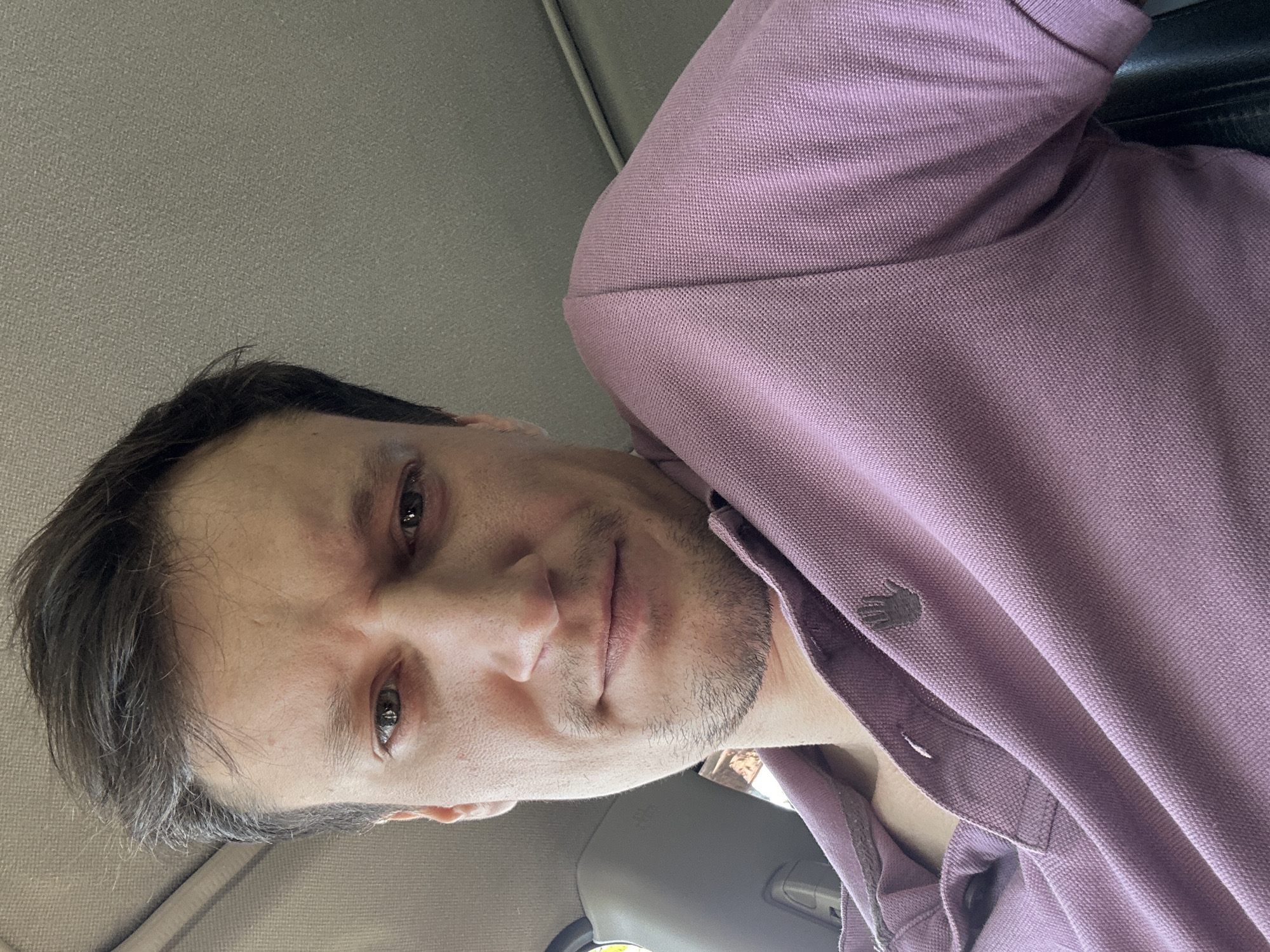 A low-angle, close-up, selfie-style photograph shows a light-skinned male with short, dark brown hair, taken inside a vehicle. The man is wearing a light purple polo shirt with a small dark logo on the left chest. His head is tilted down and slightly to the right, with his gaze directed towards the camera. His facial expression appears somber, with slightly furrowed brows and pursed lips. The background consists of the car's interior, including a gray headliner, a portion of a gray seat, and a hint of a yellow object on the left side of the frame. The overall lighting suggests it is daytime. The location is unknown.