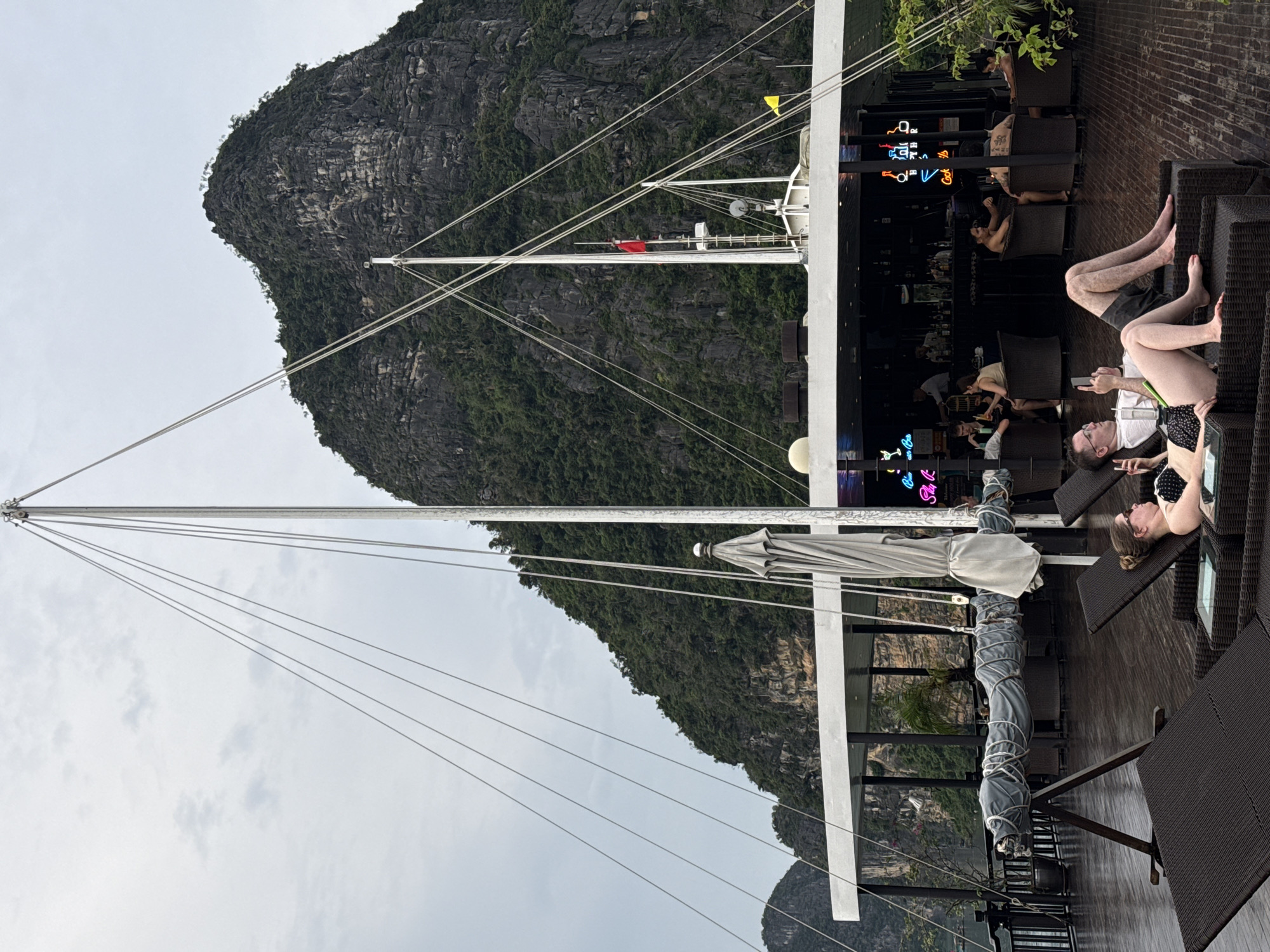 The image shows a scenic view of a boat with people relaxing on deck, with a large karst mountain in the background. The boat appears to be a tourist vessel, likely in Ha Long Bay, which is part of the larger Hải Phòng region in Vietnam. The deck is wooden and wet, suggesting recent rain. Several lounge chairs are set up, with a couple in the foreground, a woman in a black bikini and a man in a white shirt and dark shorts, both looking at their phones. Behind them, a bar area is visible with people sitting and being served. The mountain behind the boat is steep and covered in lush green vegetation. The sky is overcast.