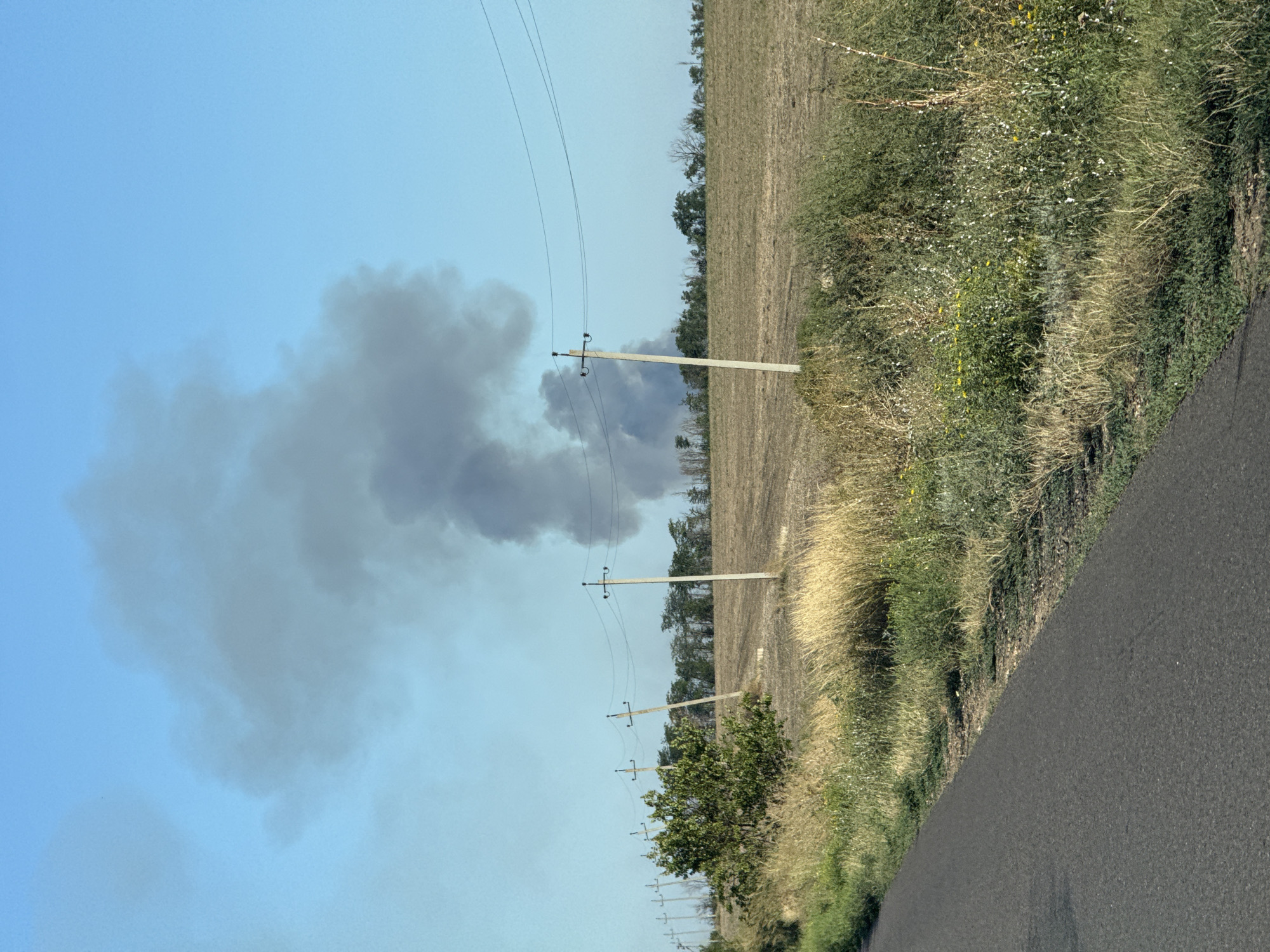 The image shows a rural landscape with a large plume of dark smoke rising into a clear blue sky. The smoke appears to be originating from a source in the distance, partially obscured by trees. In the foreground, there is a paved road running diagonally from the lower left corner. To the right of the road, there is a field with dry grass and scattered bushes. A line of utility poles with wires runs across the field. The overall impression is one of a clear day, with the dark smoke being the most prominent feature. The scene is consistent with the outskirts of Novoukrainka, Ukraine, given the rural setting and the presence of utility infrastructure.
