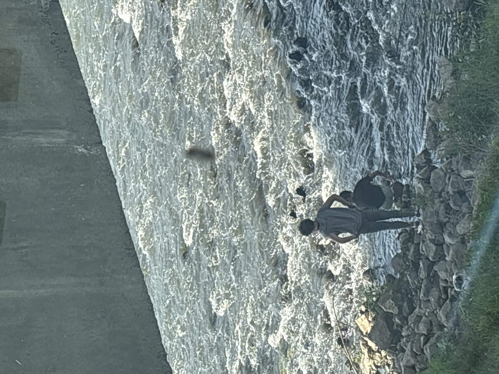 The image depicts a section of the Maritsa River in Plovdiv, Bulgaria, next to a concrete structure, possibly a bridge support or weir. Two people are standing on a rocky embankment. The river is turbulent with white water. The area is bordered by vegetation on the right.