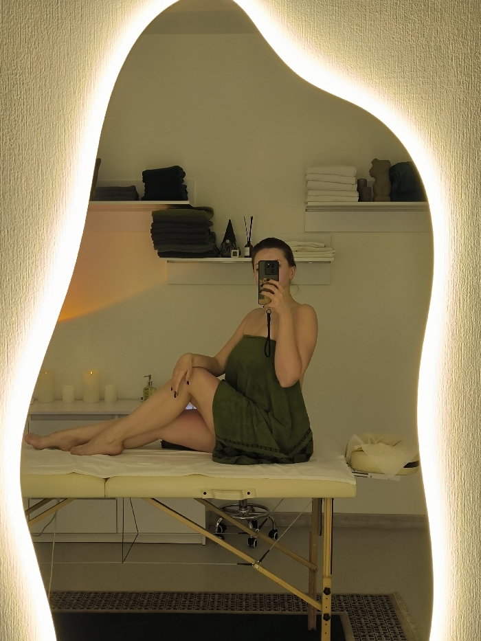 The image depicts a woman taking a mirror selfie inside what appears to be a spa or massage room in Vinnytsia, Ukraine. The woman, wrapped in an olive green towel covering her body from the chest to upper thighs, is seated on a light-colored portable massage table with a wooden frame. Her left leg is bent with her foot on the table, while her right leg is extended forward. She holds a black smartphone, obscuring her face, as she captures her reflection in an irregularly shaped, back-lit mirror that frames the scene.
The room's environment, reflected in the mirror, features minimalist white walls and warm ambient lighting from the mirror and several lit candles on a white counter to the left. Above the counter, two white wall-mounted shelves are visible. The lower shelf holds an aroma diffuser with reeds, a small dark decorative object, and a stack of white towels. The upper shelf displays stacks of dark green/grey towels on the left, more white towels on the right, and a light brown, bear-shaped decorative item. A bottle of lotion or liquid soap is present on the counter near the candles. A dark, patterned rug is partially visible on the floor beneath the massage table. The headrest of the massage table, covered with a white disposable sheet, is visible on the right side.