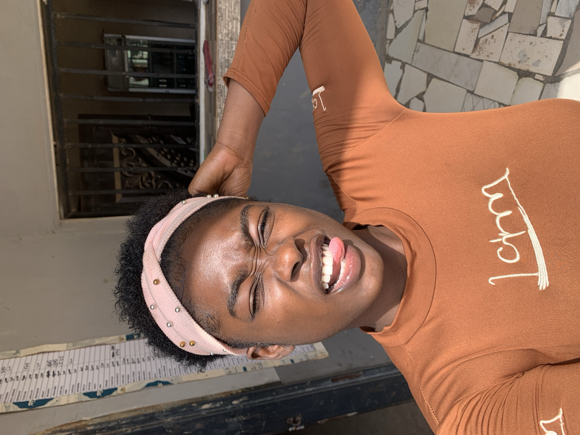 A young Black woman with dark hair and a light pink headband poses outdoors. She is wearing a brown, long-sleeved shirt with white lettering. The woman is sticking her tongue out and squinting her eyes in a playful manner.
The background includes a building with a barred window, a wall with a faded chart or sign attached, and a tiled surface. The location appears to be in Kabong, Nigeria.