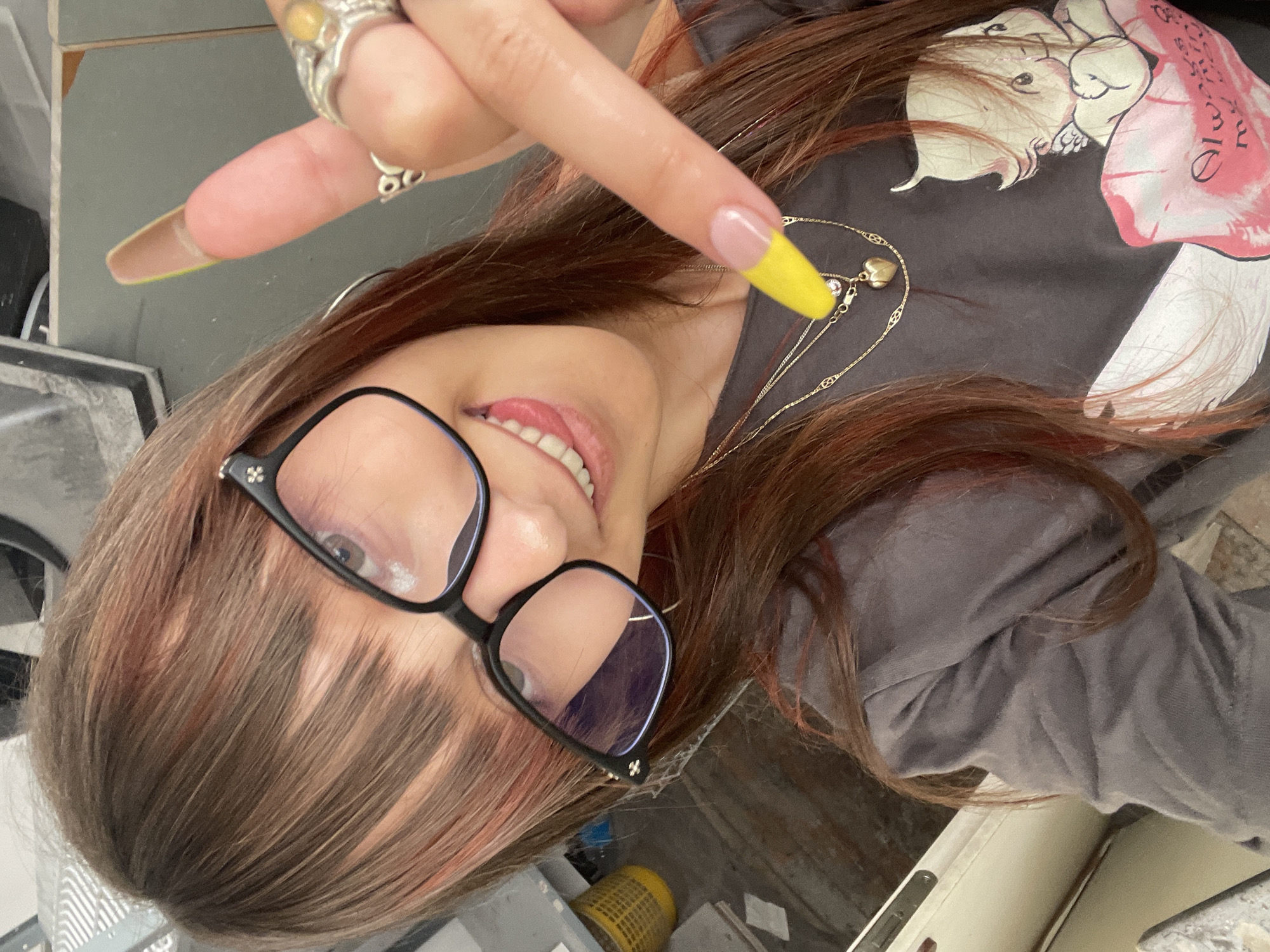 A woman, with long brown hair with reddish-pink streaks and bangs, wears large black-rimmed glasses. She is sticking her tongue out and pointing a long, bright yellow-tipped nail towards the viewer, while wearing a grey t-shirt with a graphic of a cherub holding a pink heart that says "Always mine my love". She has several necklaces on, including one with a heart pendant. The background is indistinct and appears to be an indoor or covered area, possibly a utility space or garage, with a yellow plastic basket visible to the left and a beige cabinet in the foreground. The location is identified as Raanana, Israel.