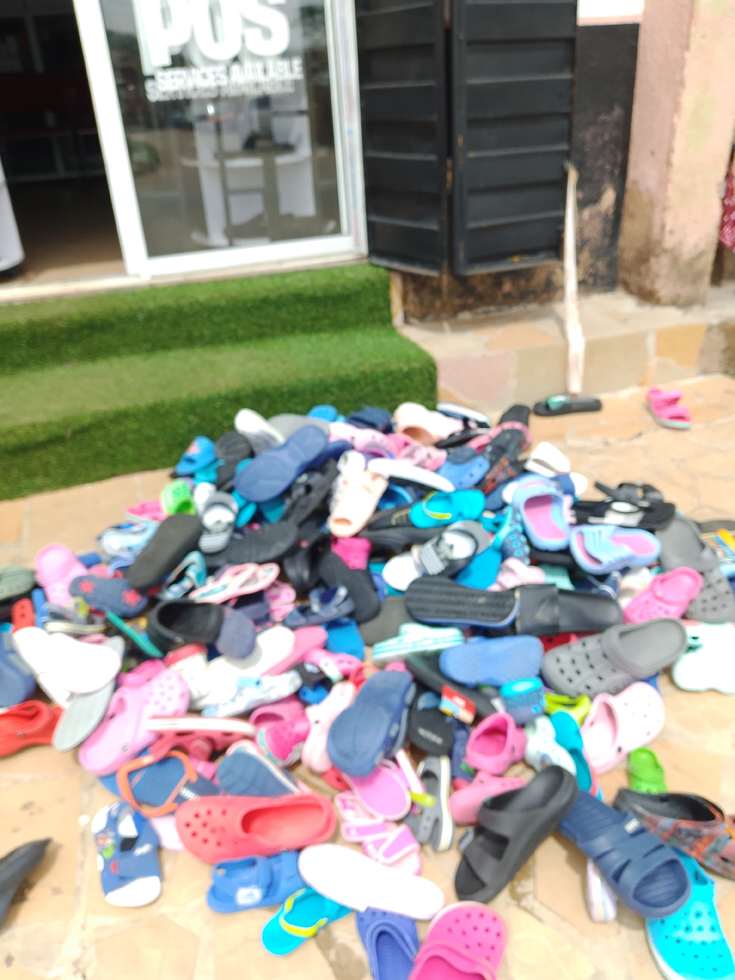 In an outdoor setting at an unknown city location, a large pile of assorted footwear, including sandals and flip-flops in various colors and styles, is displayed on the ground. The footwear is predominantly made of rubber or plastic materials. Some of the shoes are clean, while others appear to be used. The pile is situated on a textured surface, possibly pavement or stone, with a grassy area to the left and a building with black shuttered windows in the background. The lighting suggests it is daytime. There are no people visible in the image.