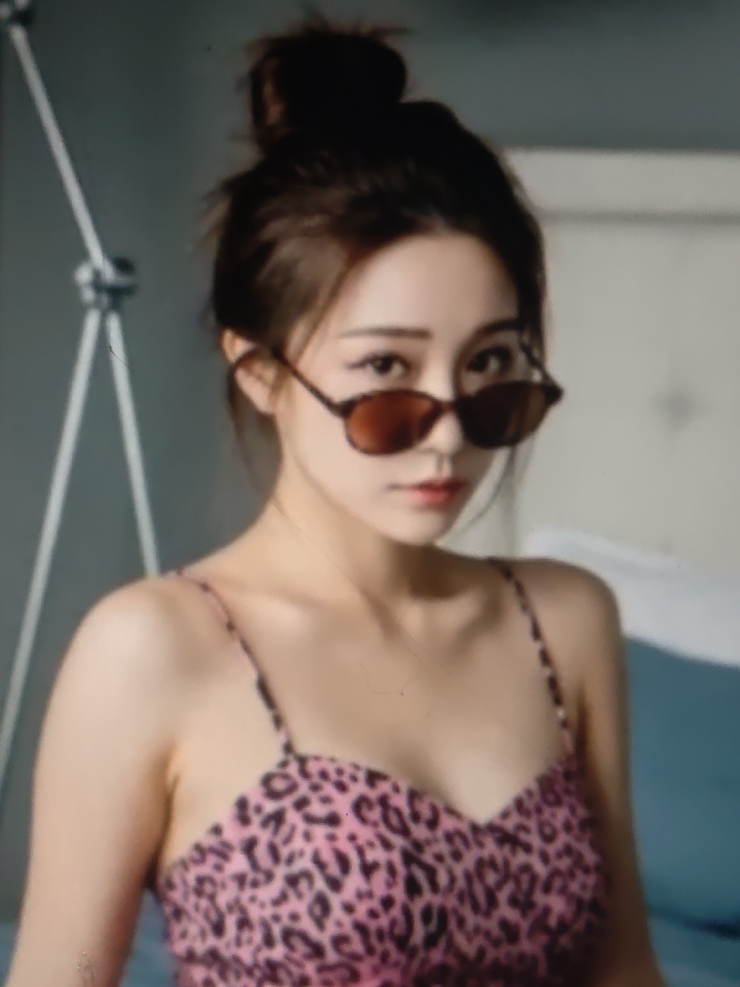 An Asian woman is posing for a picture. She has her hair in a messy bun and is wearing sunglasses and a pink leopard print camisole. She is indoors, possibly in a bedroom, with a light-colored wall behind her and a blue object to her right. There is a white tripod-like object to her left. The image is slightly blurry. There is no information in the image that would indicate the location of Kurganinsk, Russia.