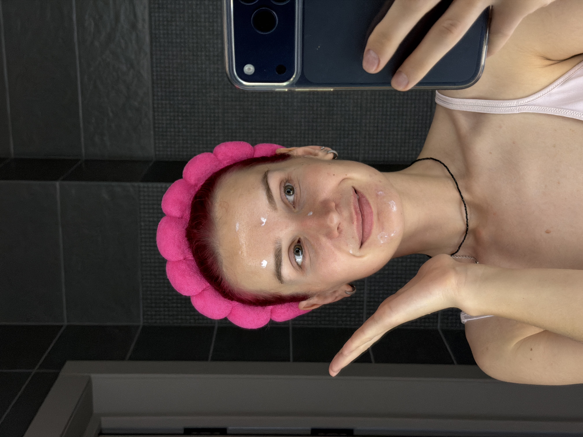 A young Caucasian woman with short, bright red hair, wearing a bright pink fluffy headband and a light pink camisole, is taking a selfie in what appears to be a bathroom. She has a few white dabs of cream on her face, particularly on her forehead, cheeks, and chin. Her left hand is raised and positioned near her face. She is looking at her phone, which she is holding in her right hand. The background shows dark, textured tiles on the wall and a sliver of a white door or frame on the left. The lighting is somewhat dim, with some light reflecting off her skin and the phone screen. There is no indication of the city of Chaikovsky, Russia, in the image.