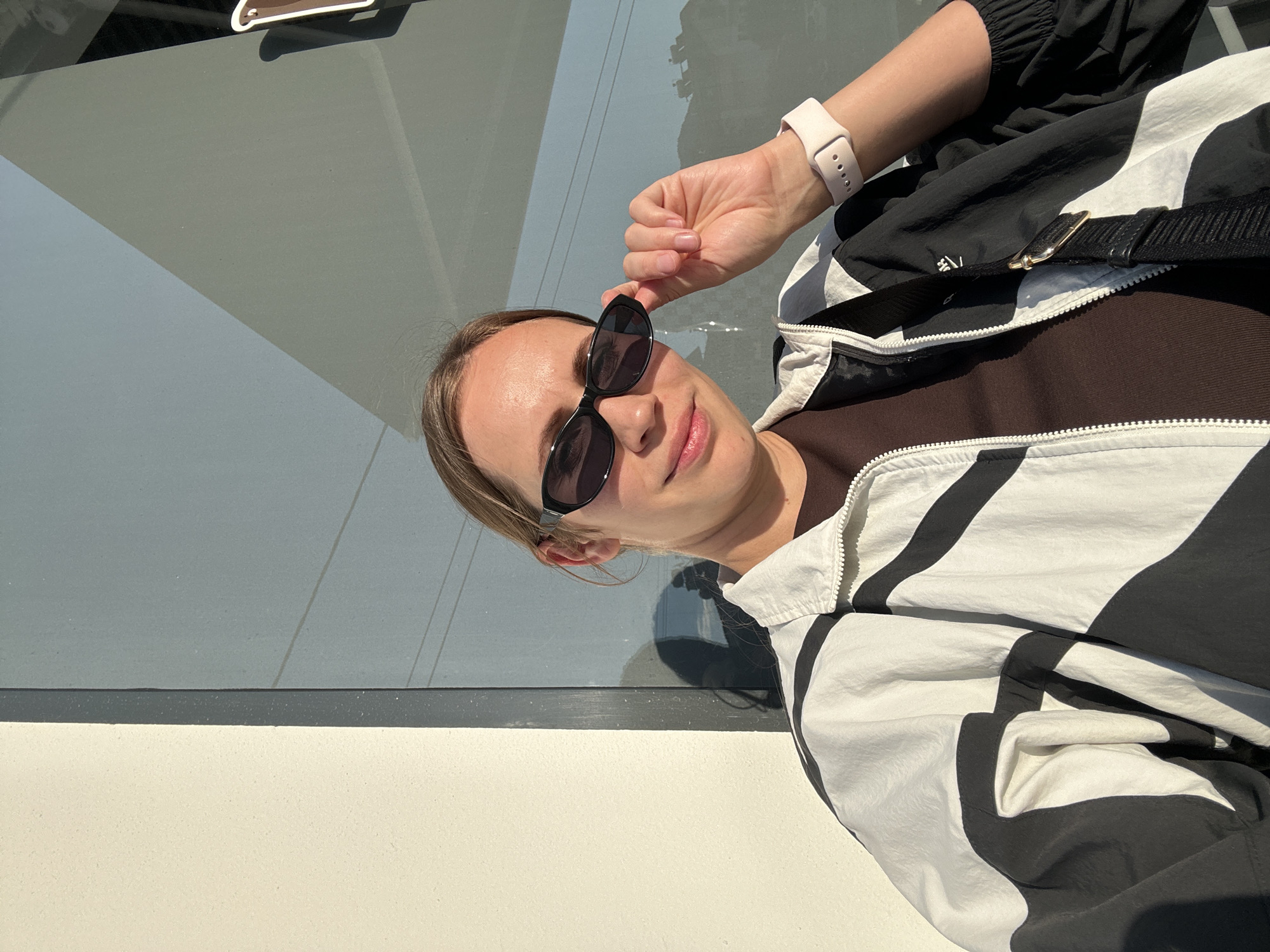 A fair-skinned woman is posing for a selfie outdoors on a sunny day. She is wearing sunglasses, a brown top, and a black and white patterned jacket. She has a light-colored smartwatch on her left wrist. A black strap is visible across her chest. The background includes a building with a glass window and a white wall. The location is likely in the city of Fethiye, Turkey, based on the request.