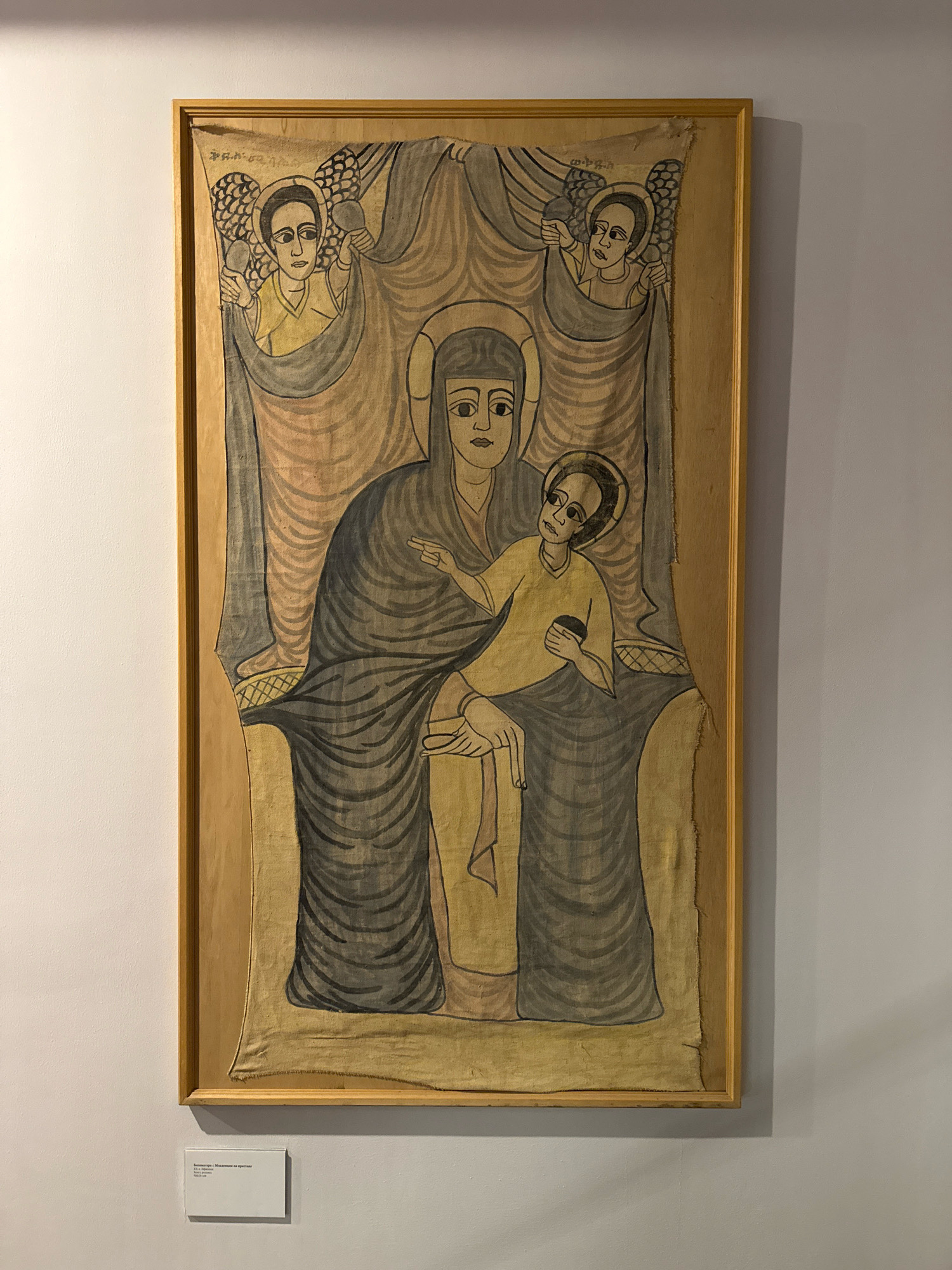 The image shows a framed Ethiopian Orthodox painting of the Virgin Mary and Child, flanked by two angels, hanging on a white wall in a museum. The painting is on fabric and appears to be old, with some fraying around the edges. The figures are rendered in a stylized, two-dimensional manner typical of Ethiopian art, with large eyes and simple clothing. The Virgin Mary wears a dark blue robe, and the Child, depicted as a young boy, is dressed in yellow. The angels also have stylized wings and halos. A small white plaque is visible below the painting on the wall, likely providing information about the artwork. The overall lighting is even, suggesting an indoor exhibition space. The painting is centrally positioned on the wall, with ample white space surrounding it.