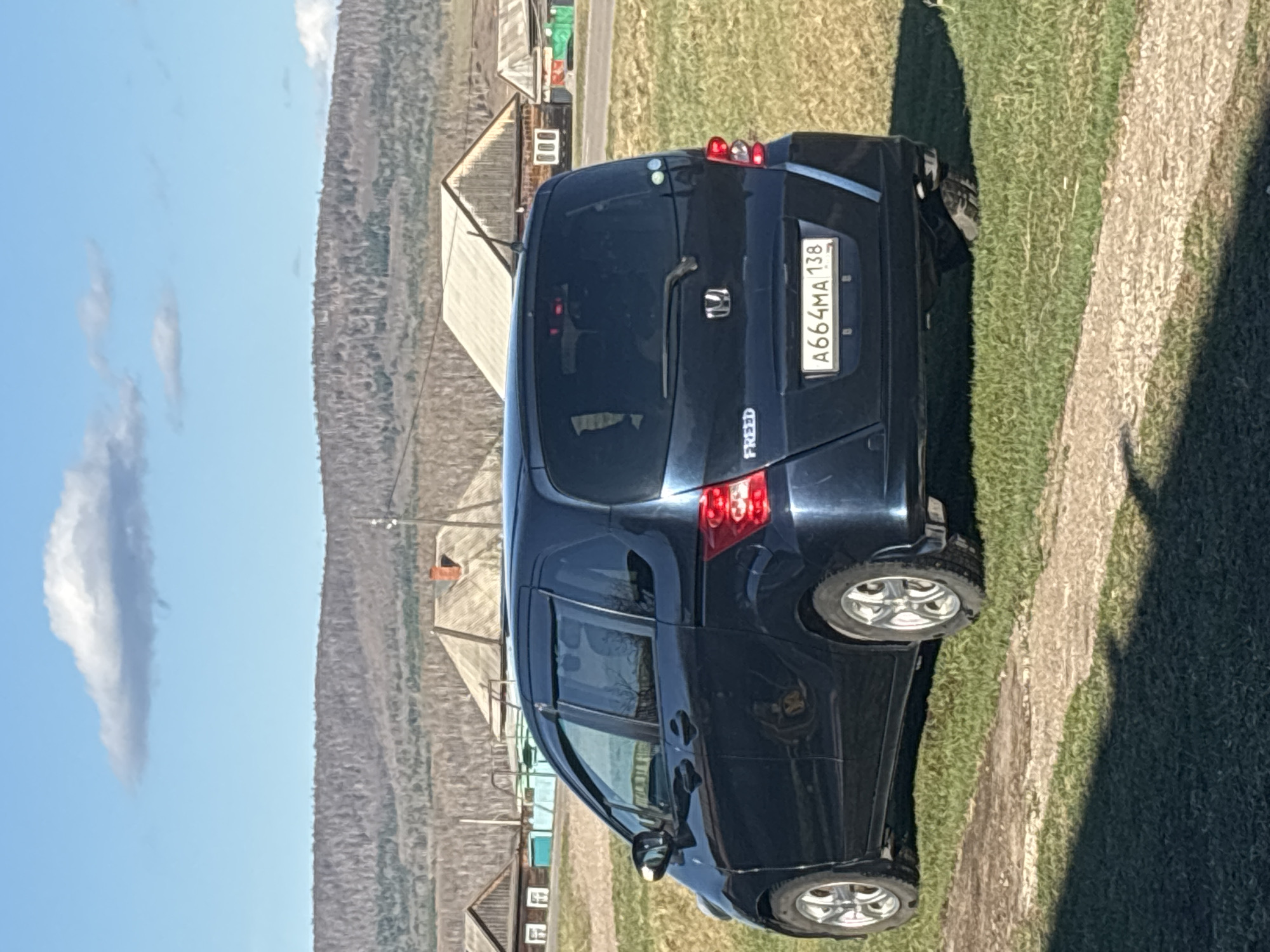 The image shows a dark blue Honda Freed minivan parked on a grassy area next to a dirt road. The vehicle's rear is facing the viewer, with the license plate A664MA 138 visible. In the background, there are several wooden houses, typical of rural architecture, and a treed hillside. The sky is blue with some clouds. Based on the license plate region code "138," the image is likely taken in the Zabaykalsky Krai region of Russia. The presence of rustic wooden houses suggests a location in a small village. Given the provided location of "Zagatuy, Russia," it is likely the image was taken in or around that city. There are no people visible in the image.