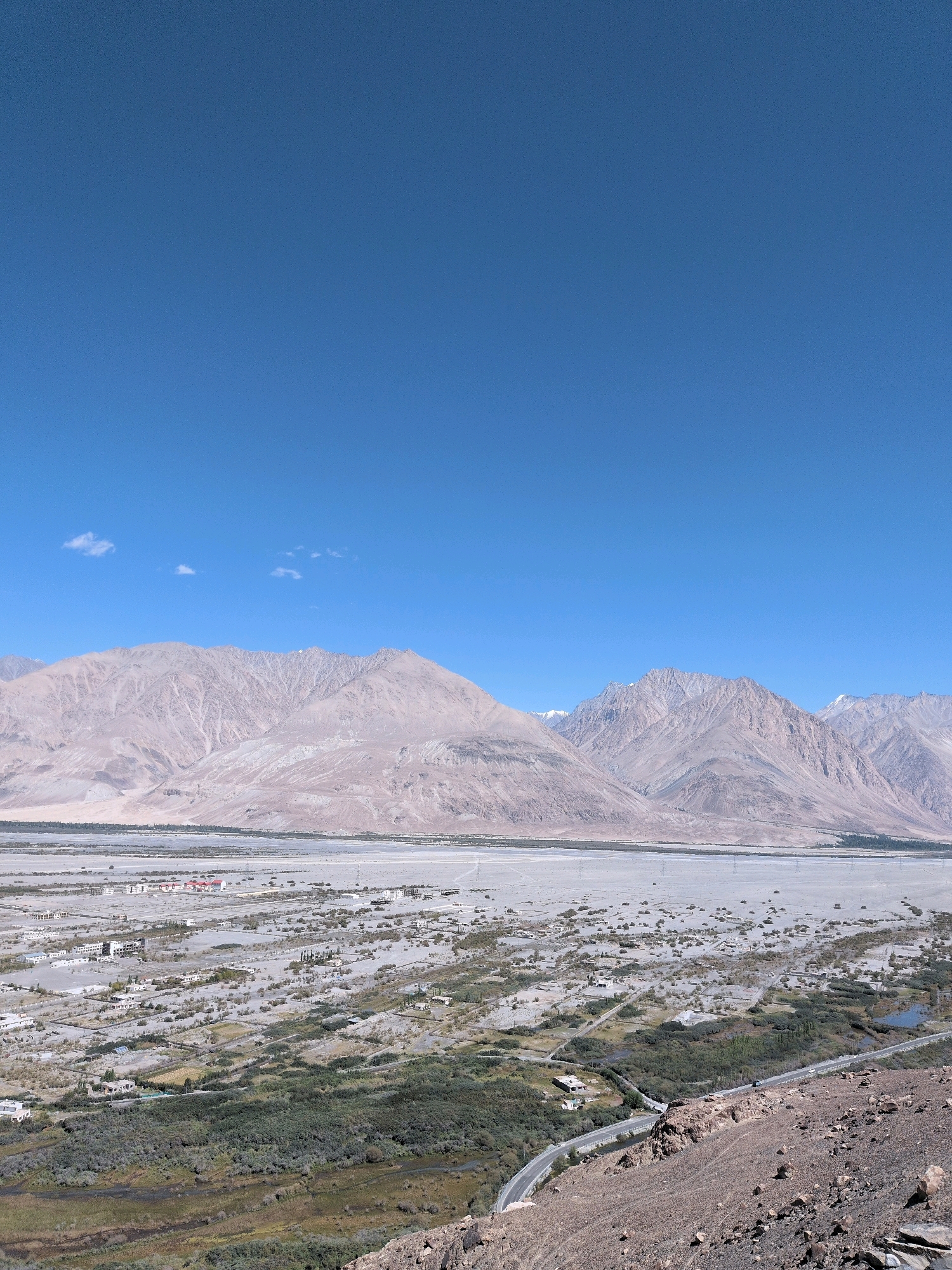 This image captures a panoramic view of the city of Diskit, India, under a clear blue sky. The scene is dominated by the stark beauty of the landscape, with towering, arid mountains forming a dramatic backdrop. In the foreground, a winding road is visible, traversing a rugged, rocky terrain. Beyond the road, a green, vegetated area with a small body of water gives way to the more sparsely populated outskirts of Diskit. The city itself appears as a collection of buildings spread across a vast, dusty plain, with some structures clustered together and others more isolated. The overall impression is one of a remote, high-altitude settlement nestled within a grand, natural environment. There are no people or active interactions visible in the image.