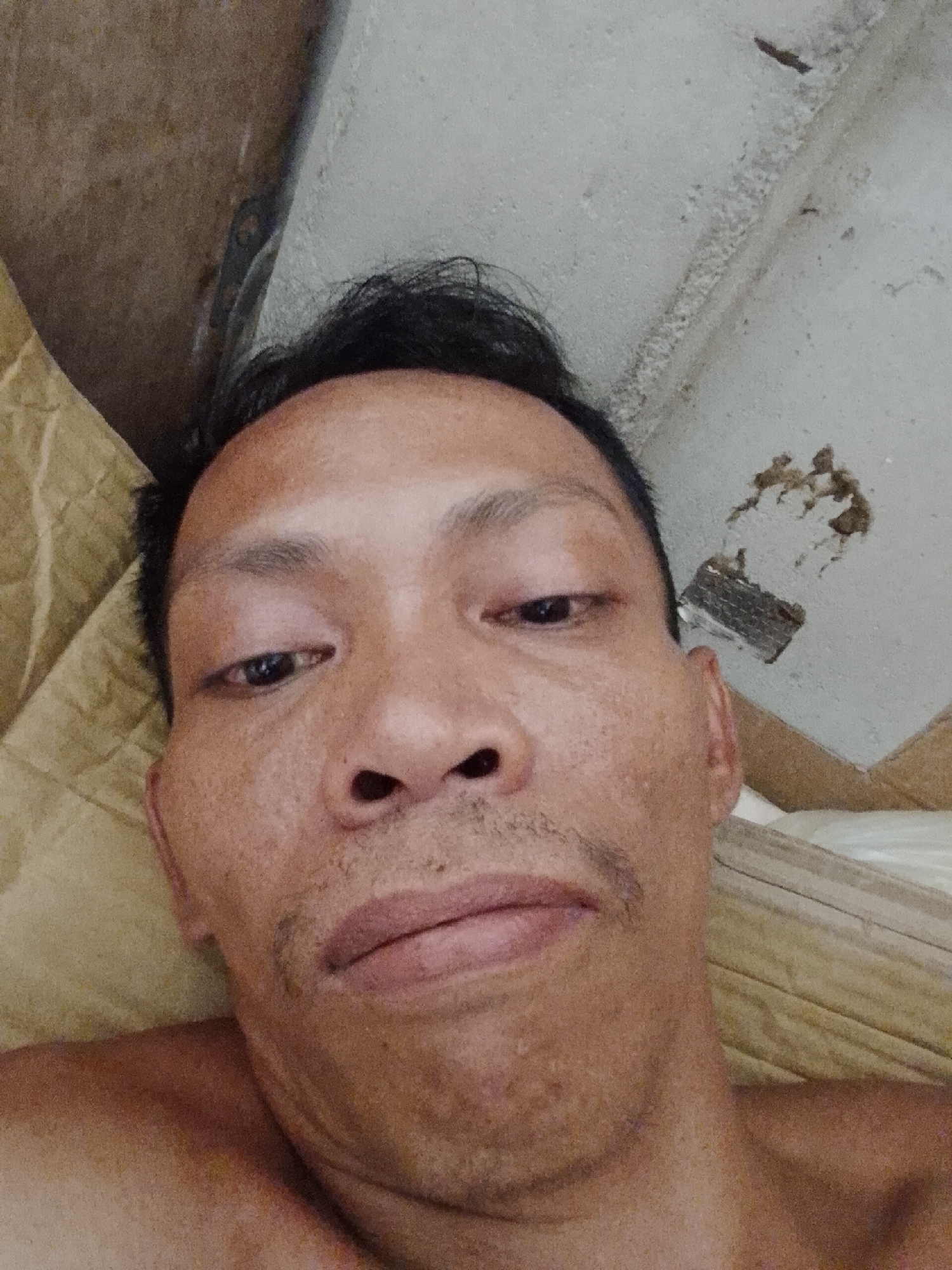 The image is a close-up selfie of a man with dark hair. He appears to be lying down, with a tan quilted surface behind him and a rough, grey ceiling above. The ceiling has some damage and exposed material. The man has dark eyes, a mustache, and appears to have skin imperfections. The setting is unknown without more context. No information indicates that this image was taken in Bukit Ketok, Indonesia.