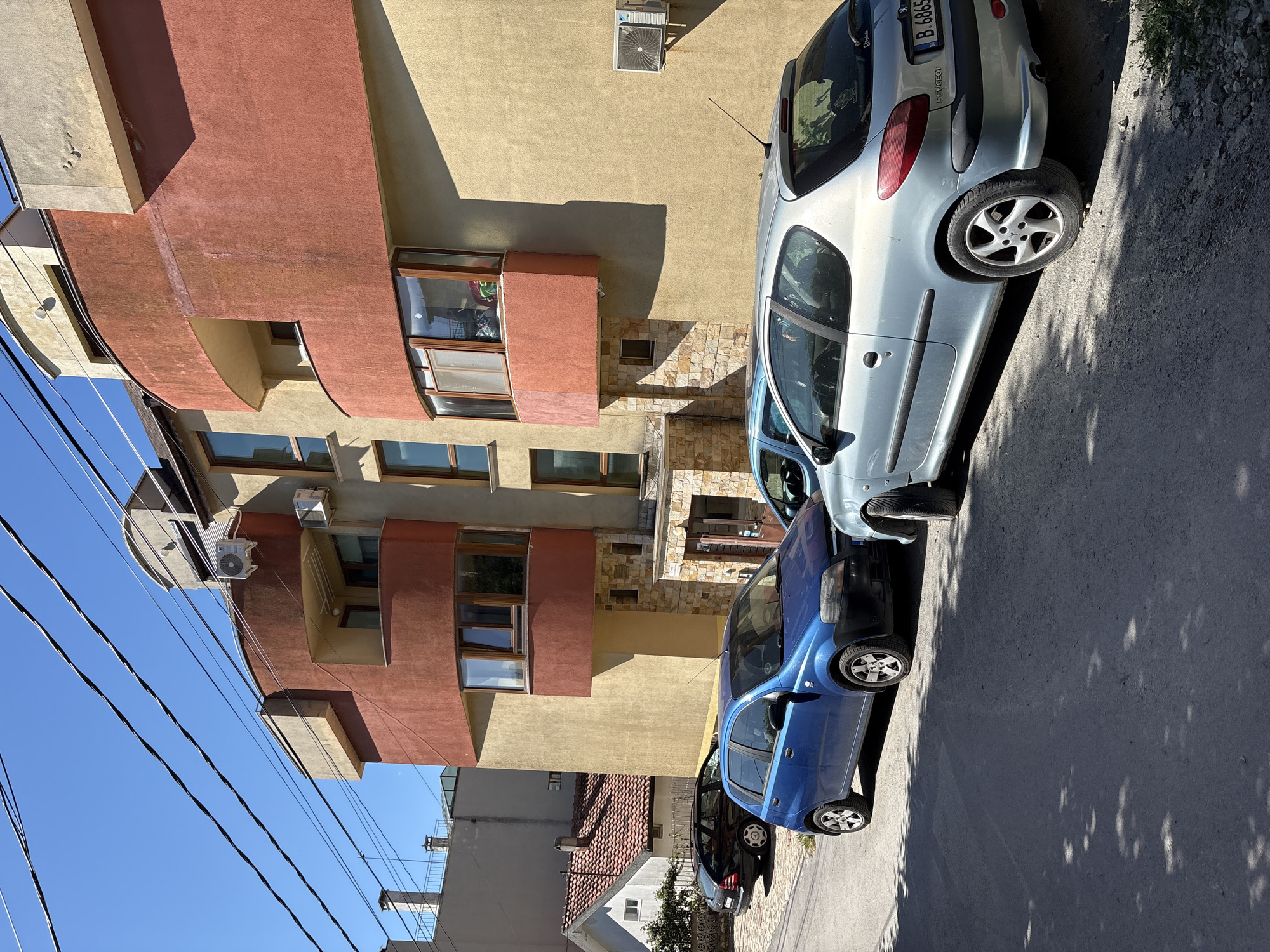 The image shows a street scene in Varna, Bulgaria, with a modern apartment building dominating the background. The building features a mix of yellow and reddish-orange stucco facades, with curved balconies and large windows. Several cars are parked on the paved street in front of the building.
In the foreground, a light blue Peugeot 206 is parked facing away from the camera. Its rear license plate is visible and reads "B 6865 BD". Next to it, a darker blue Chevrolet Aveo hatchback is parked, facing the same direction. Further down the street, a black Mercedes-Benz sedan is parked.
The sky is clear and blue, indicating a sunny day. Overhead power lines are visible, crossing the frame. To the left of the apartment building, a smaller, older house with a red-tiled roof is partially visible. The street appears to be a quiet residential area.
The scene is static, with no people visible in the frame. The cars are parked, and the environment is typical of a European city street. The overall impression is one of everyday urban life.