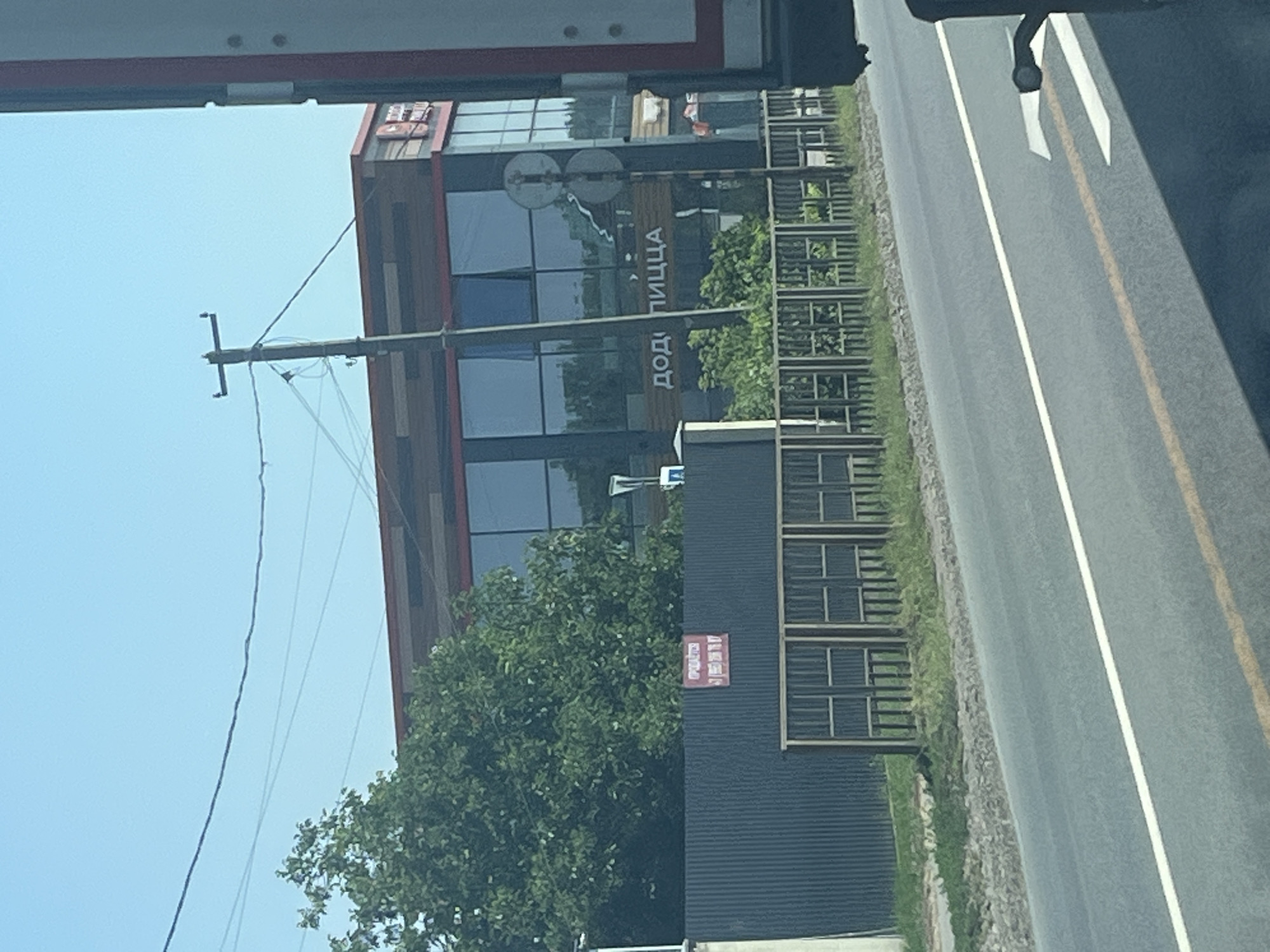 An eye-level, medium shot captures a commercial building with a modern design, featuring a dark wood-paneled facade with large glass windows. The words "ДОДО ПИЦЦА" are visible in large white letters on a wooden sign on the building. A tall utility pole with wires extends upwards into the clear blue sky. In front of the building, a dark corrugated metal fence stands, with a smaller wooden fence in front of it. Green trees are visible behind the fence, and lush green grass grows along the side of a paved road in the foreground. Part of a white truck is visible on the right side of the frame, obscuring some of the building. The overall scene appears to be a street view of a commercial establishment.