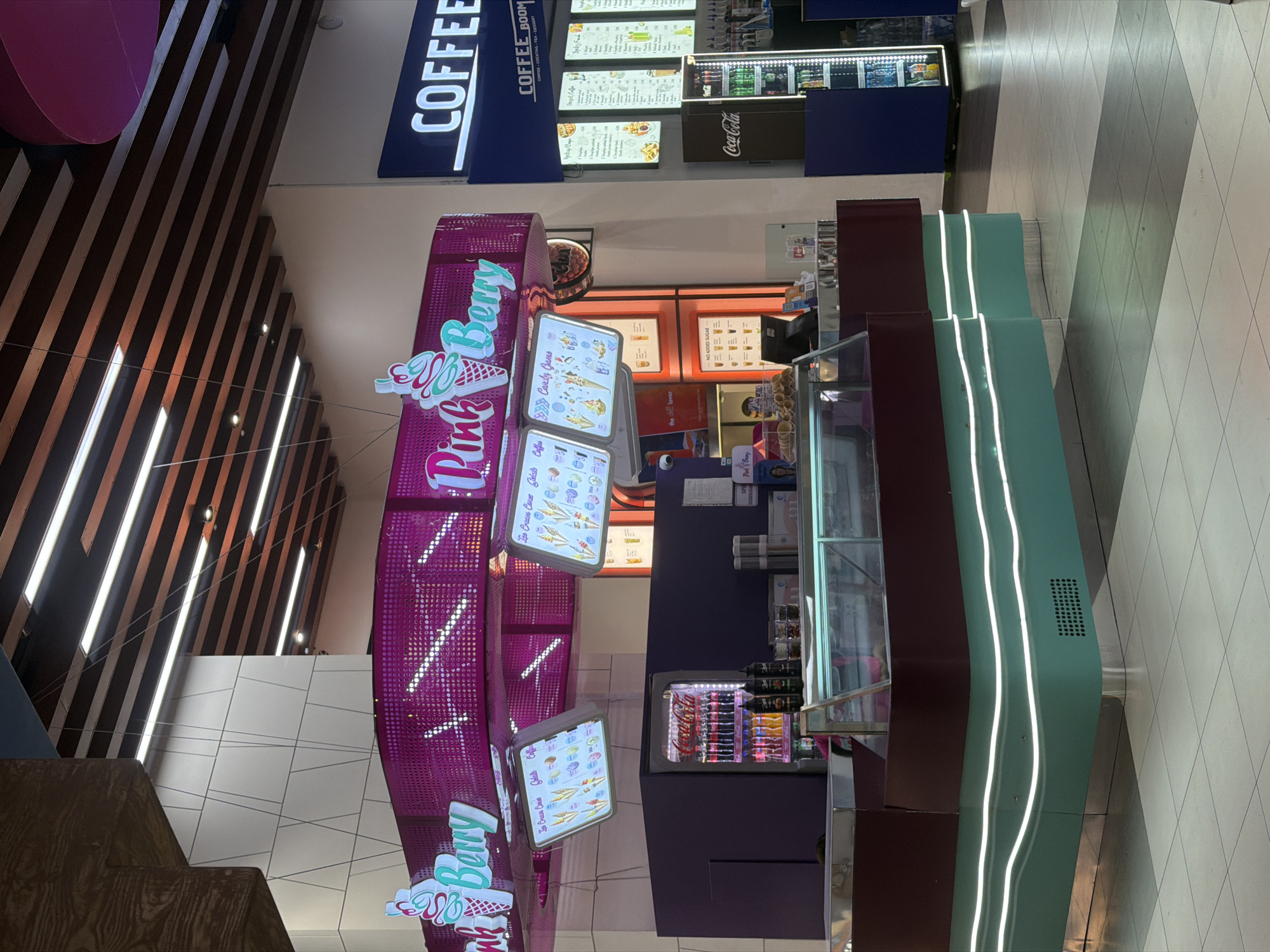 In Yerevan, Armenia, within an indoor commercial setting, a Pink Berry ice cream and dessert shop is depicted. The shop features a vibrant pink canopy adorned with lights and the shop's name in stylized lettering, accompanied by ice cream cone graphics. Below the canopy, illuminated menu boards display various ice cream creations, gelato, and coffee options. A refrigerated display case showcases what appears to be ice cream. To the right of the Pink Berry shop, a "COFFEE BOOM" establishment is visible, with its own illuminated menu boards offering coffee, cocktails, tea, and desserts. A Coca-Cola branded refrigerator is situated next to the coffee shop. The environment appears to be a mall or a similar indoor public space, with tiled flooring and a modern architectural design for the ceiling and surrounding structures. No people are clearly visible in the scene.