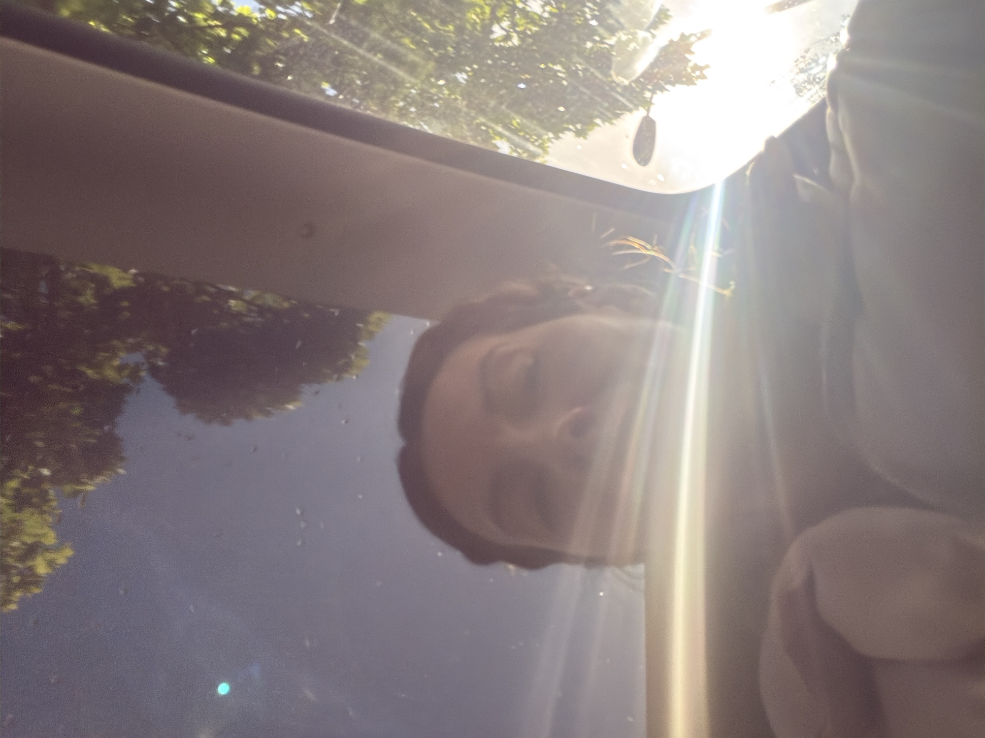 The image shows a person inside a car, likely seated in the driver's or passenger's seat, looking up through the sunroof. The person has their eyes closed. Through the sunroof, the sky and treetops are visible. The reflection on the glass suggests it is a bright, possibly sunny, day. The location is not identifiable as London, United Kingdom.