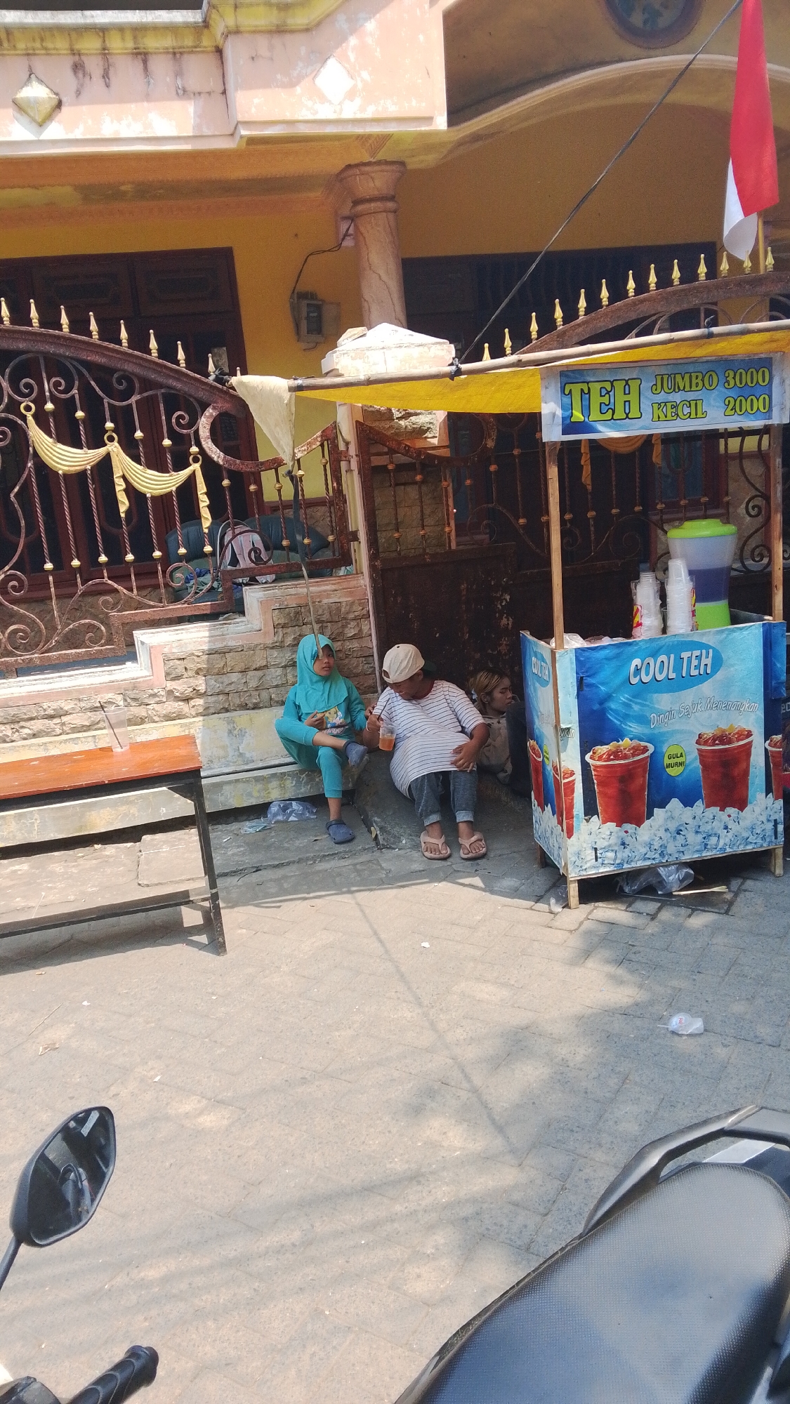 In Mancar, Indonesia, a street scene unfolds with a small beverage stall and individuals sitting nearby. The stall, labeled "COOL TEH," features an image of iced tea and is adorned with a yellow tarp. Two people are seated on the ground next to the stall, one a person wearing a blue hijab and a teal outfit, holding a cup. Next to them, a person in a striped t-shirt and cap drinks from a similar cup. Further back, another individual is partially visible. The stall is situated on a paved area, with a metal fence and a building in the background. A wooden table is placed to the left of the scene. The overall environment suggests a warm day, with shadows cast by overhead structures.