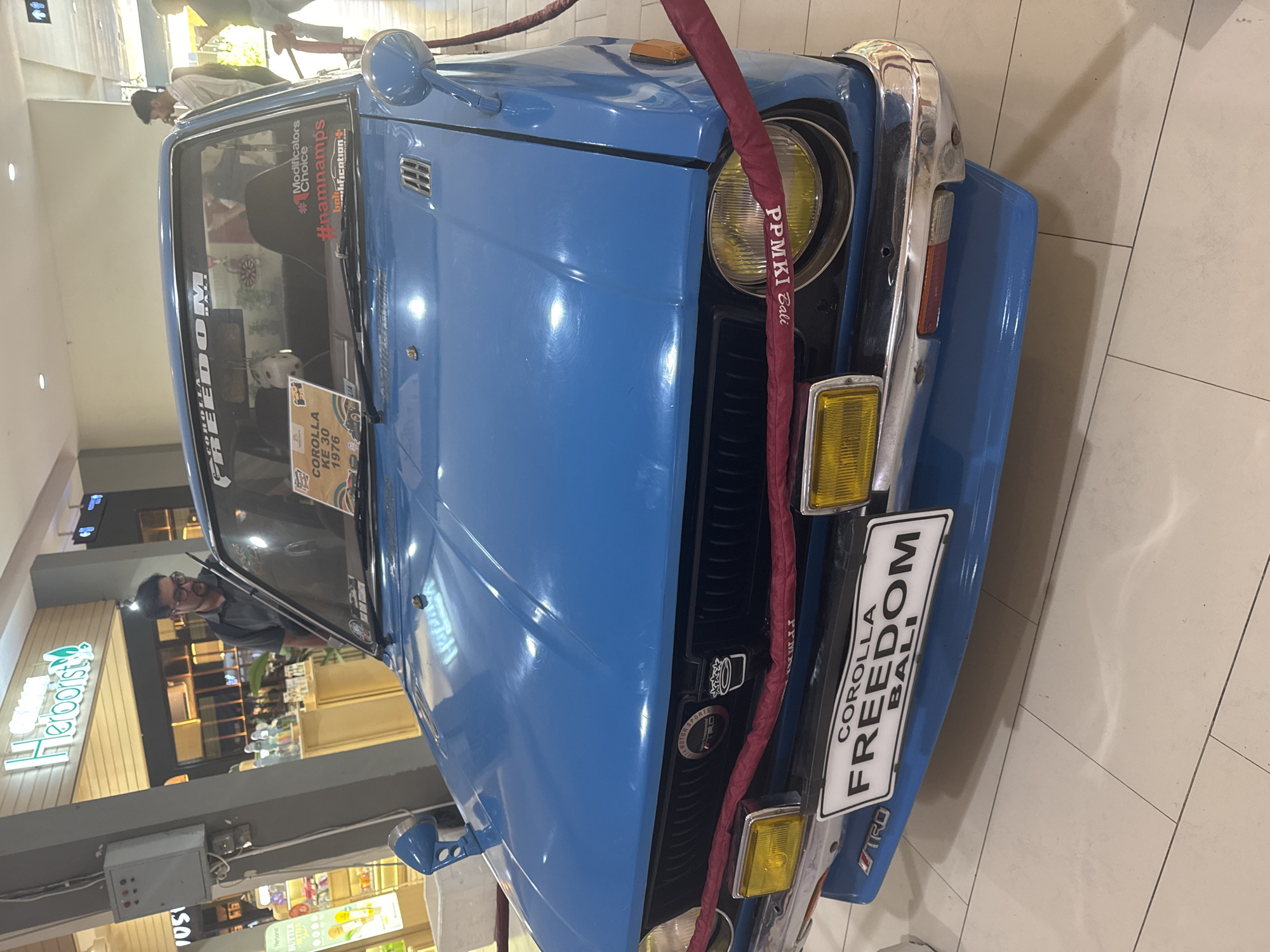 The image shows a blue, vintage car, identified by its license plate as a "COROLLA FREEDOM BALI." A sign on the windshield indicates it is a "COROLLA KE 30 1976." The car is prominently displayed indoors, likely in a shopping mall or exhibition hall, as suggested by the tile floor and the presence of shops in the background, including one named "Herborist." A burgundy protective cover is draped around the front. There are people visible in the reflections on the car's windshield. A light fixture is visible in the top of the image.