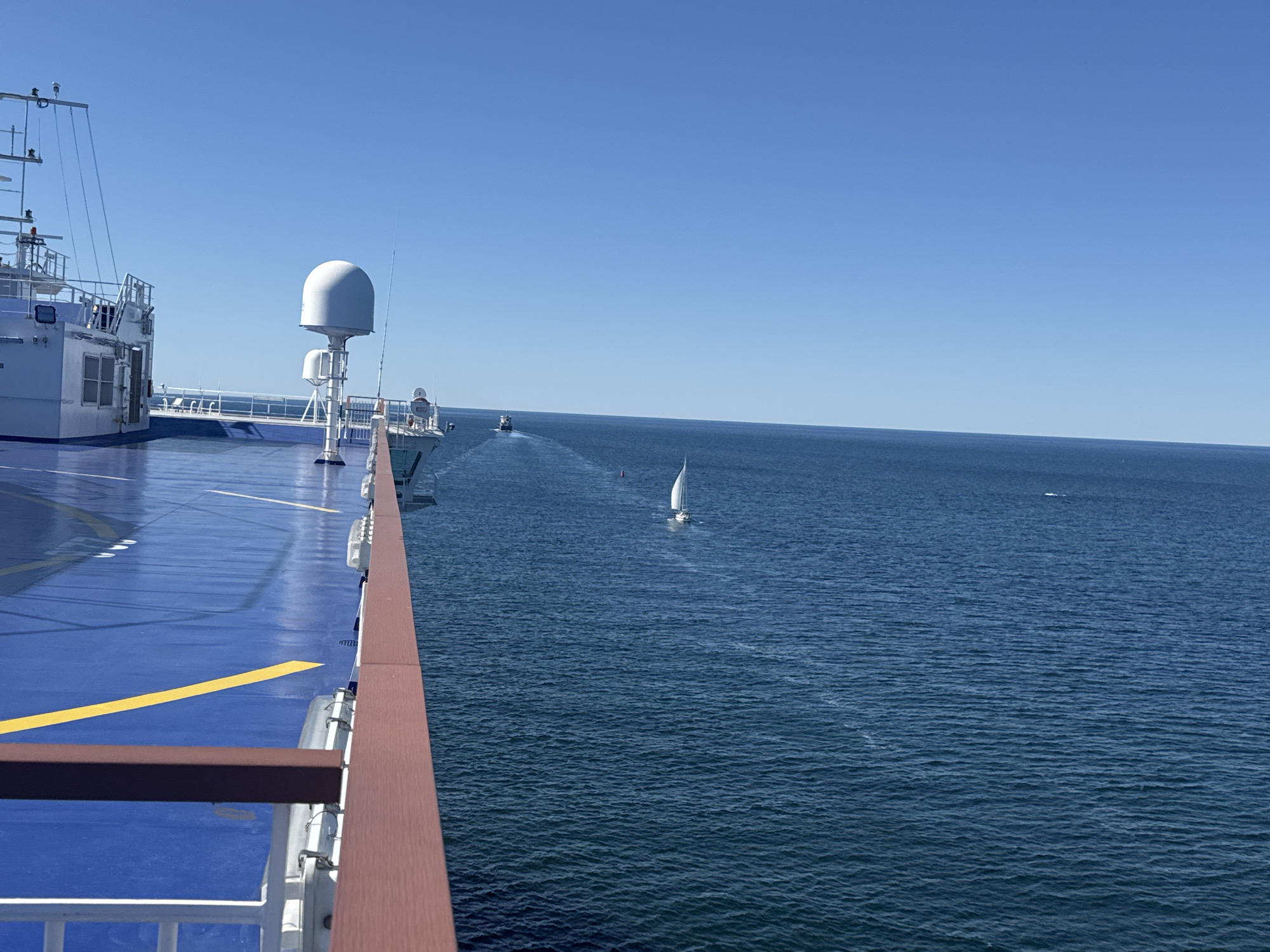 The original image shows a view from a large white ferry, likely the Stena Danica, with its blue deck and yellow lane markings visible in the foreground. The ferry is at sea under a clear blue sky. In the distance, a small white sailboat is visible, navigating the deep blue ocean. Further in the distance, a smaller boat leaves a wake behind it, also heading towards the horizon. The location is implied to be near Drottningskär, Sweden, suggesting the ferry is traversing the waters of the Baltic Sea. There are no people visible in the image. The overall scene is one of a calm, sunny day at sea.